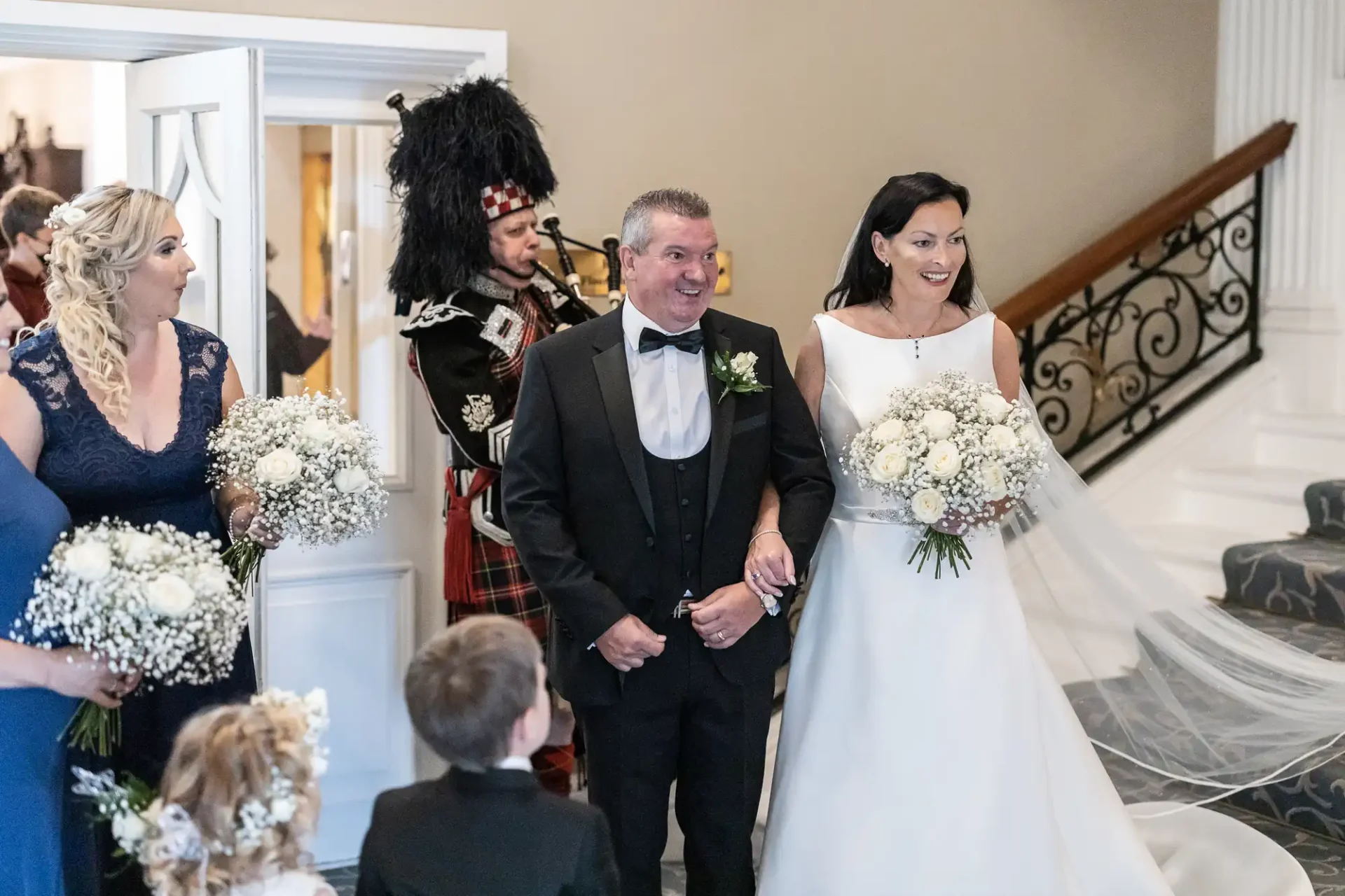 A bride and groom walk down stairs at their wedding, accompanied by a bagpiper in traditional scottish attire, with guests looking on.