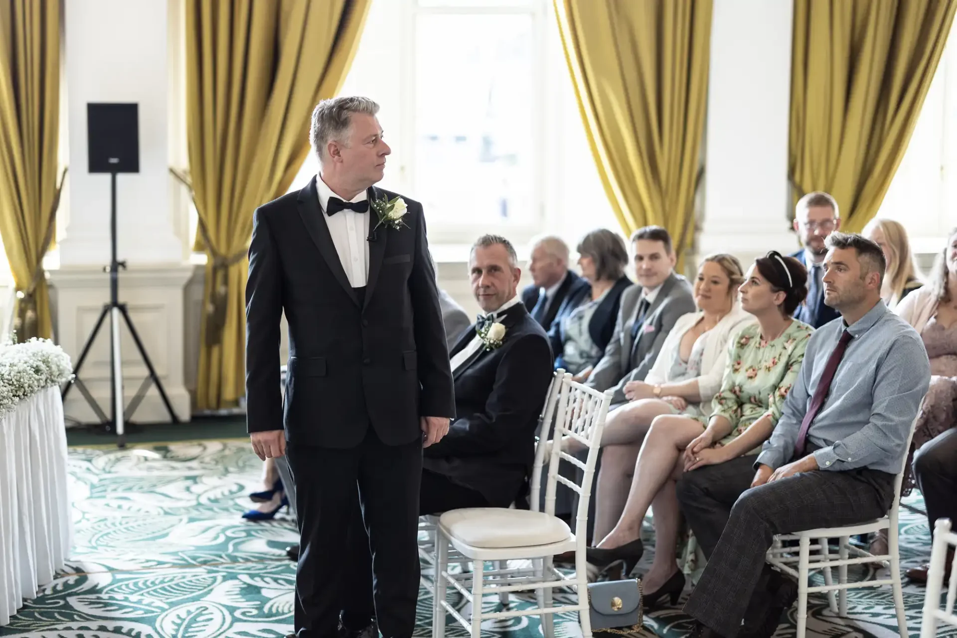 A groom in a black tuxedo stands at the front of a wedding ceremony in an elegant room, with seated guests looking on.