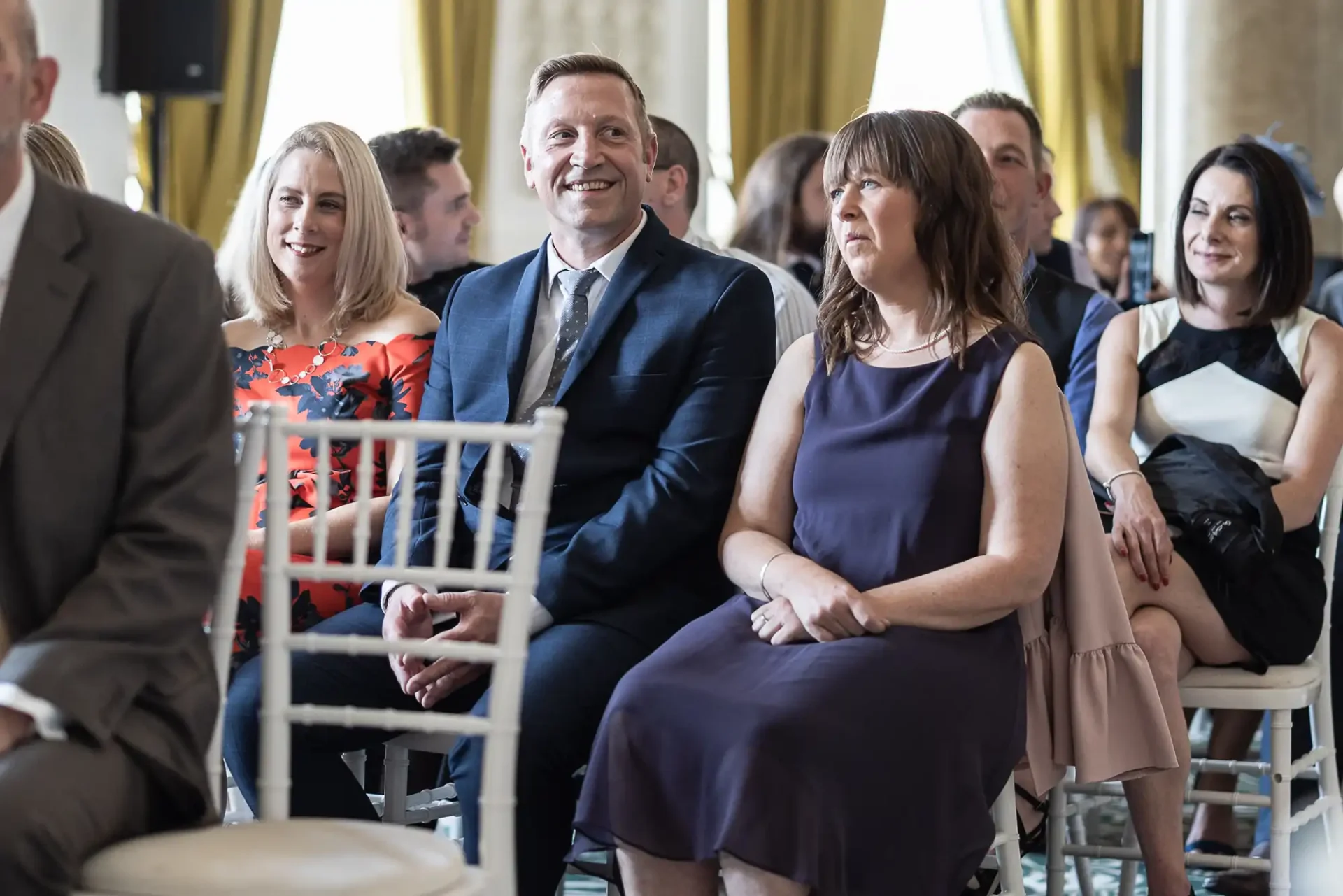 Audience members seated at an event, smiling and focused on the front, in a venue with elegant decor.