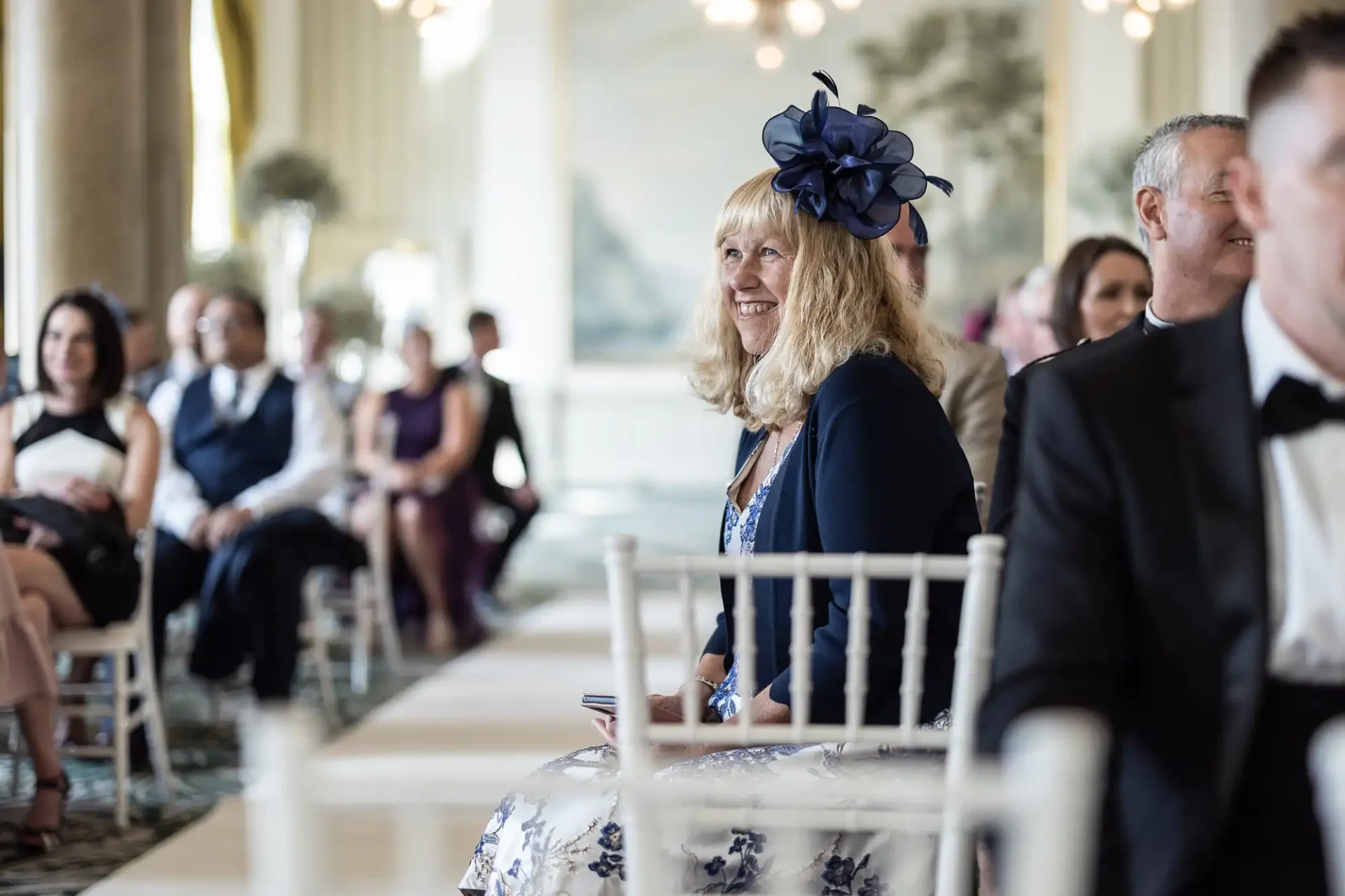 Woman in a blue hat and dress smiles while seated at an elegant wedding ceremony in a lavishly decorated room.