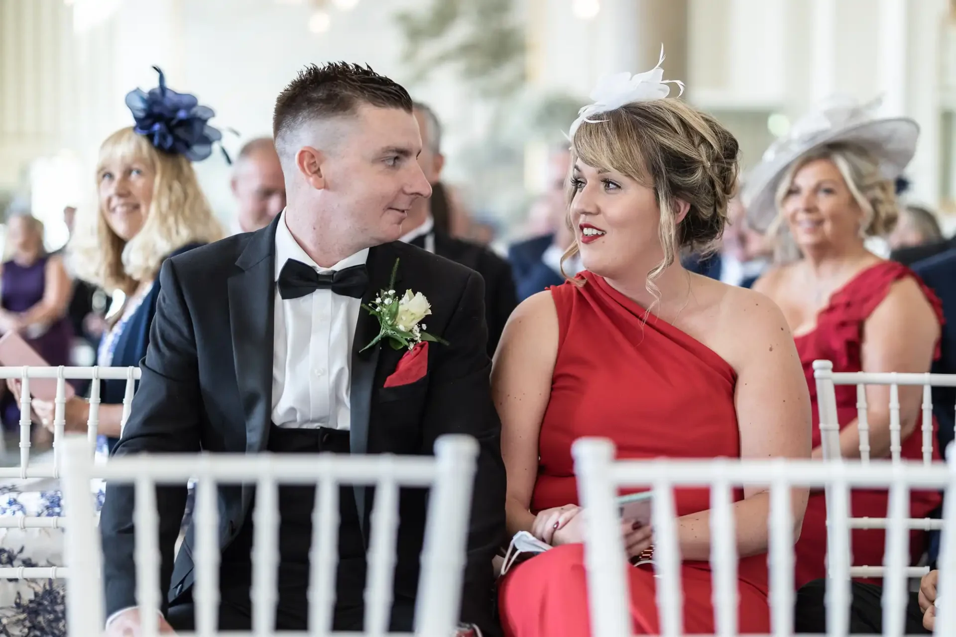 A couple dressed in formal wear sitting at a wedding ceremony, the man in a black tuxedo and woman in a red dress, both looking at each other smiling.