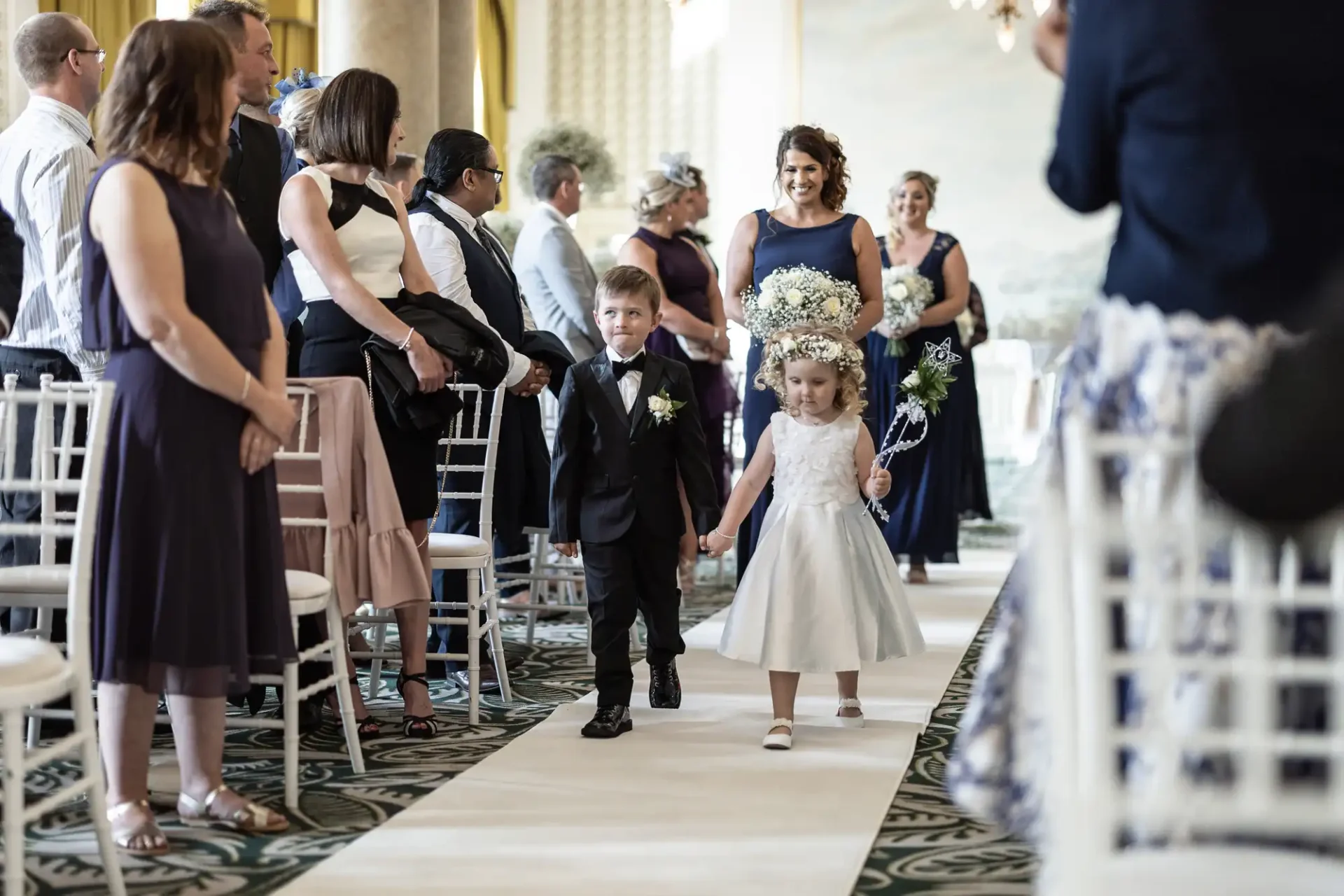 A young boy in a suit and a young girl in a white dress walking down the aisle at a wedding, with guests watching and smiling.