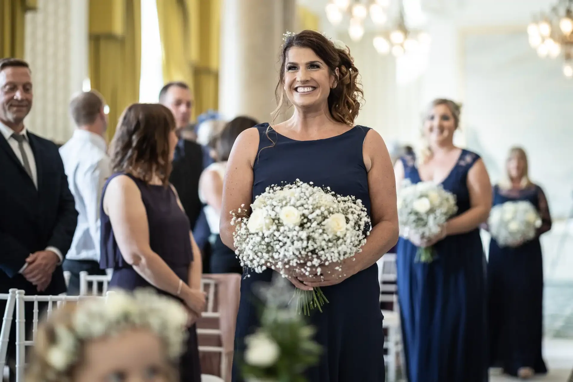 A woman in a blue dress smiling while holding a bouquet of white flowers at a wedding ceremony, with guests in the background.