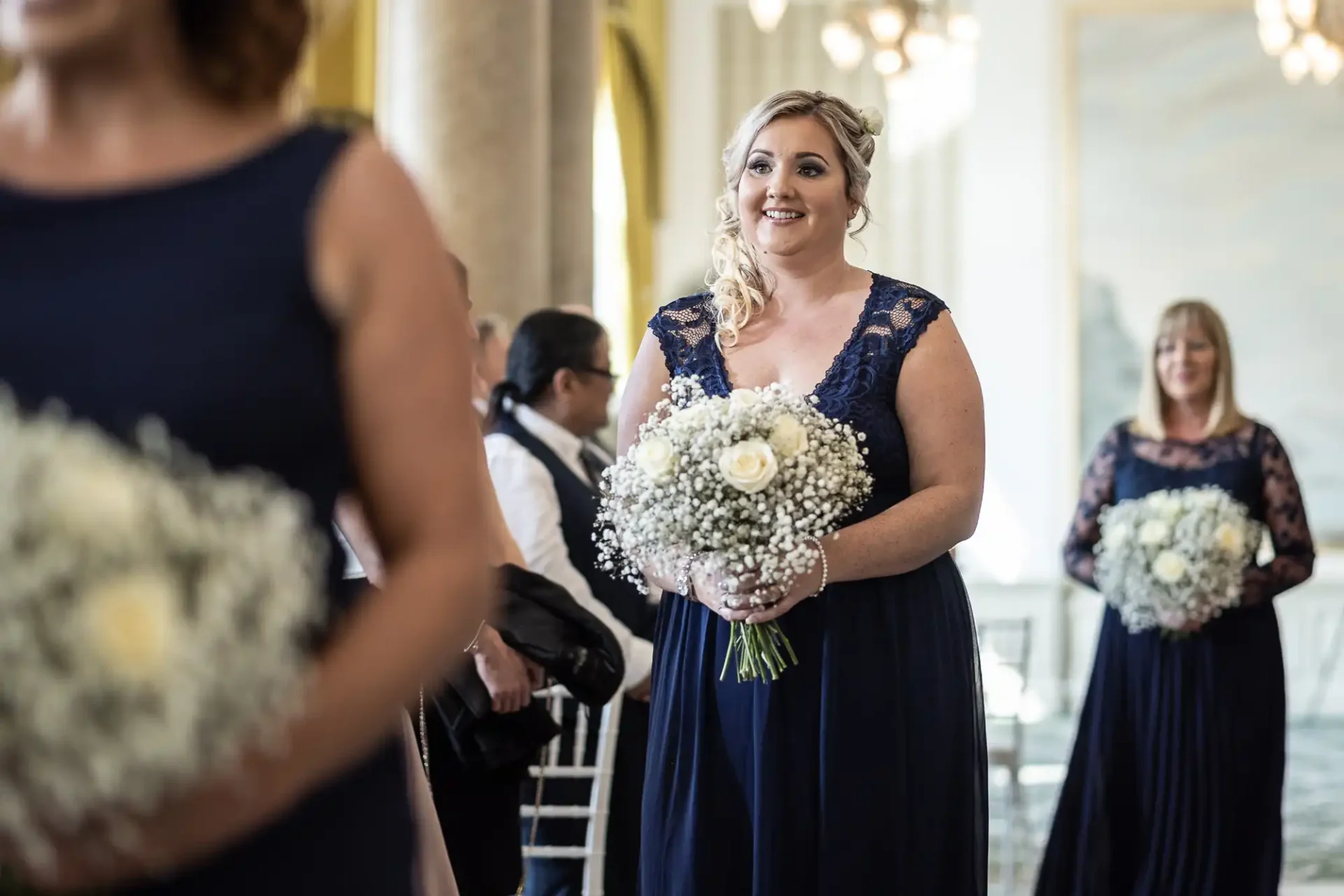 A woman in a navy blue dress smiles, holding a bouquet, at a wedding ceremony with other guests around.