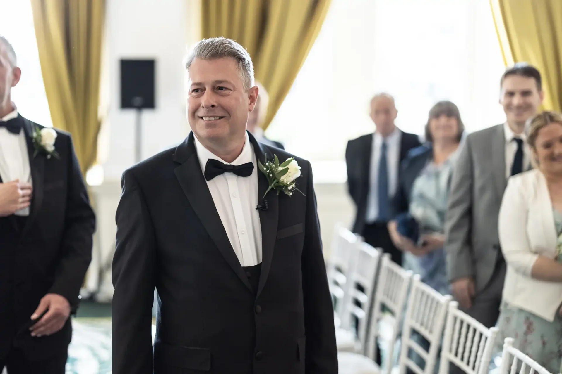 A smiling man in a tuxedo with a boutonniere stands at a wedding ceremony, surrounded by guests in a well-lit room with white chairs.