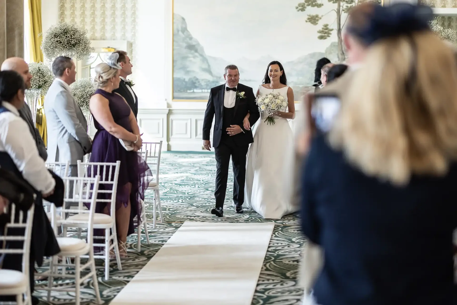 A bride walking down the aisle with her father, guests watching on either side in a decorated room.