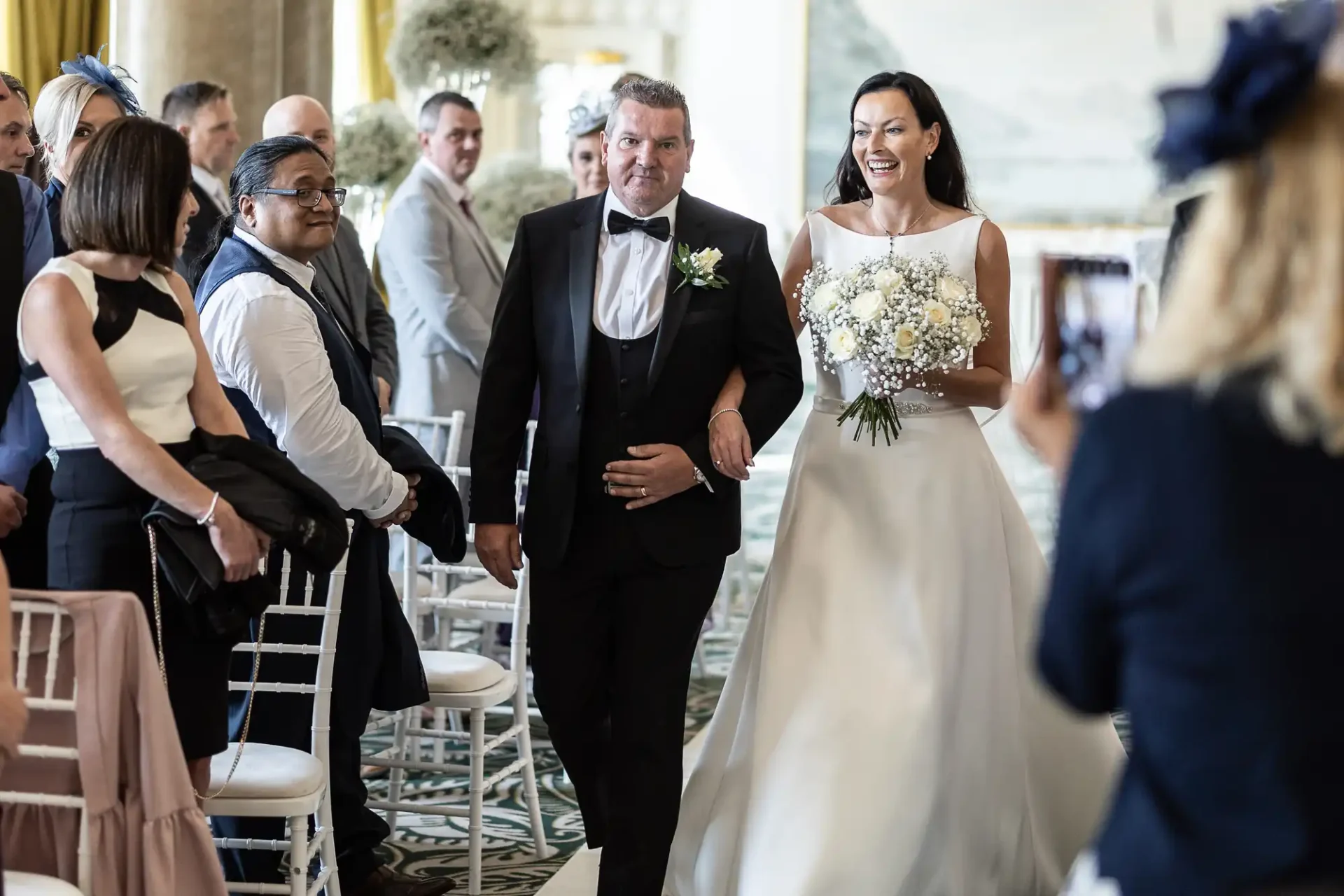 A bride and groom walking down the aisle, smiling, as guests watch and one guest takes a photo.
