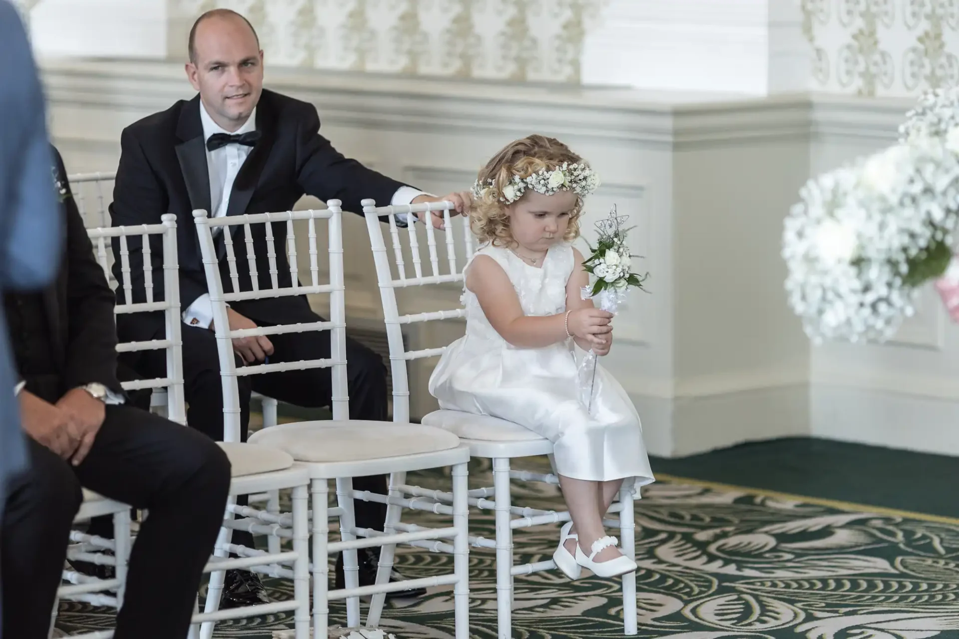 A man in a tuxedo sits beside a young girl in a white dress and floral headband, who is holding a small bouquet, both seated at a formal event.