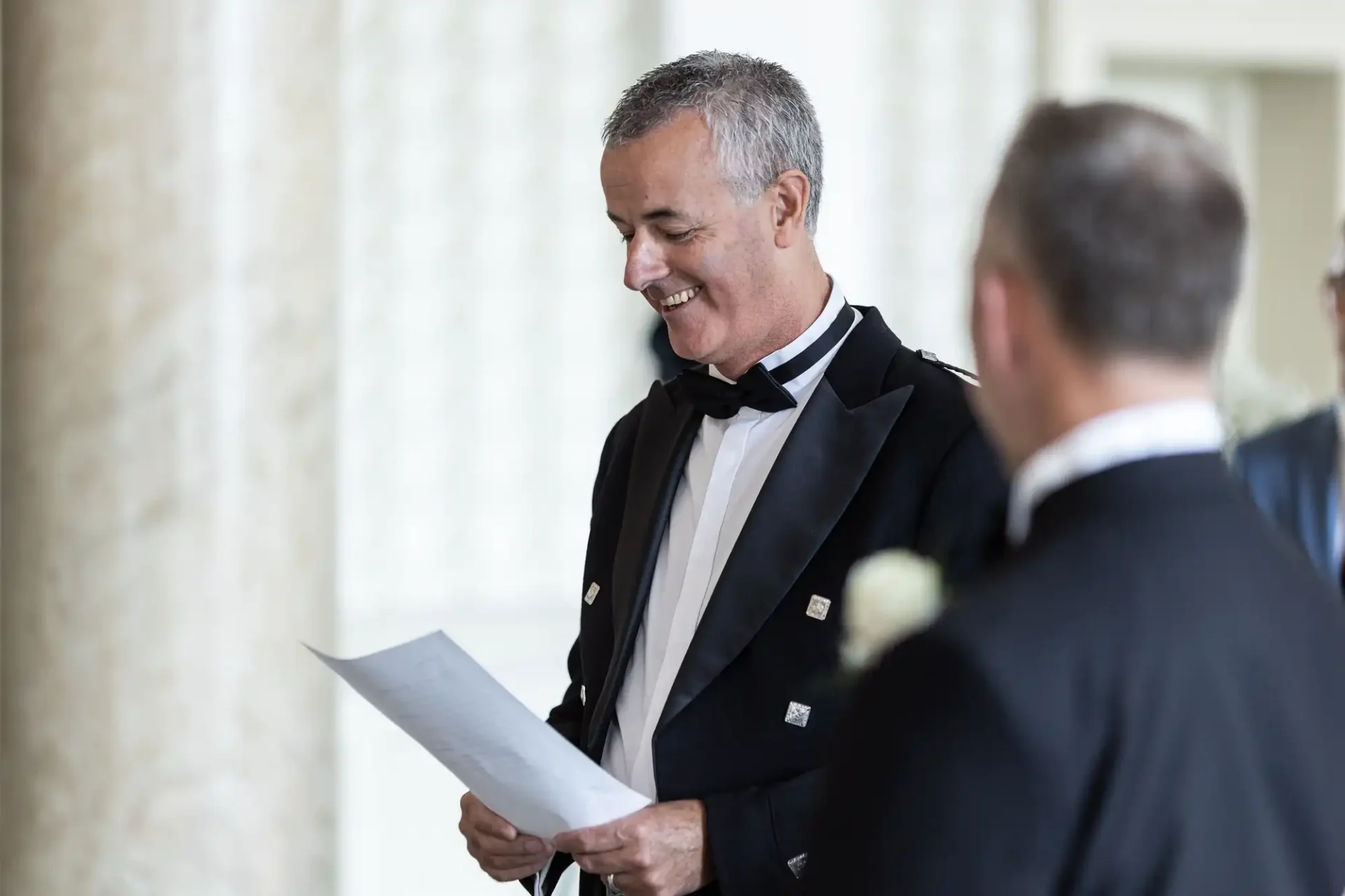 A smiling man in a tuxedo with a bow tie reading a document, standing next to another man in a similar outfit at an indoor event.
