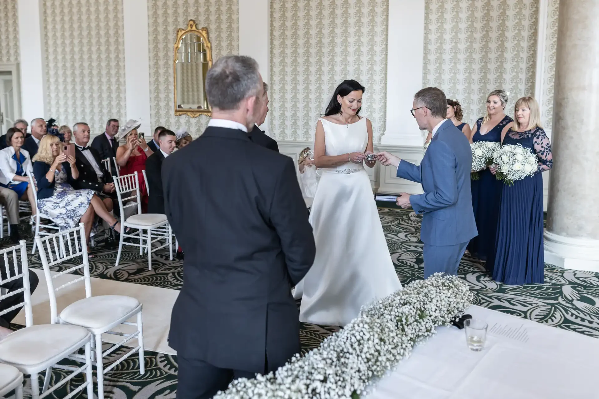 A bride and groom exchange rings during their wedding ceremony in a grand room filled with guests.