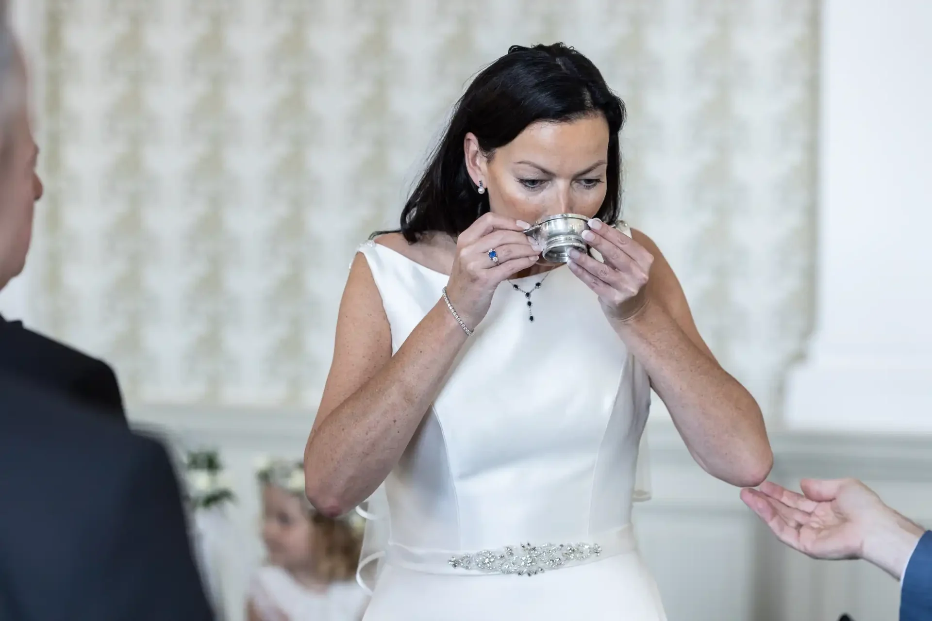 A bride in a white dress sipping from a small silver cup during a wedding ceremony, with a blurred background showing a young girl.