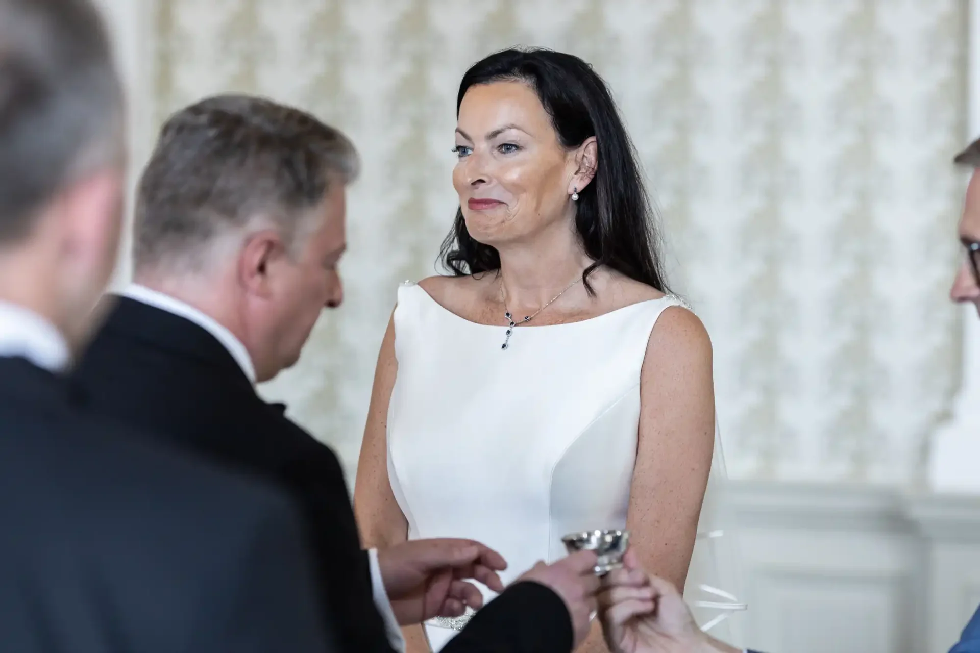 A woman in a white dress smiles while interacting with a man in a suit at a formal event, with another attendee partially visible.