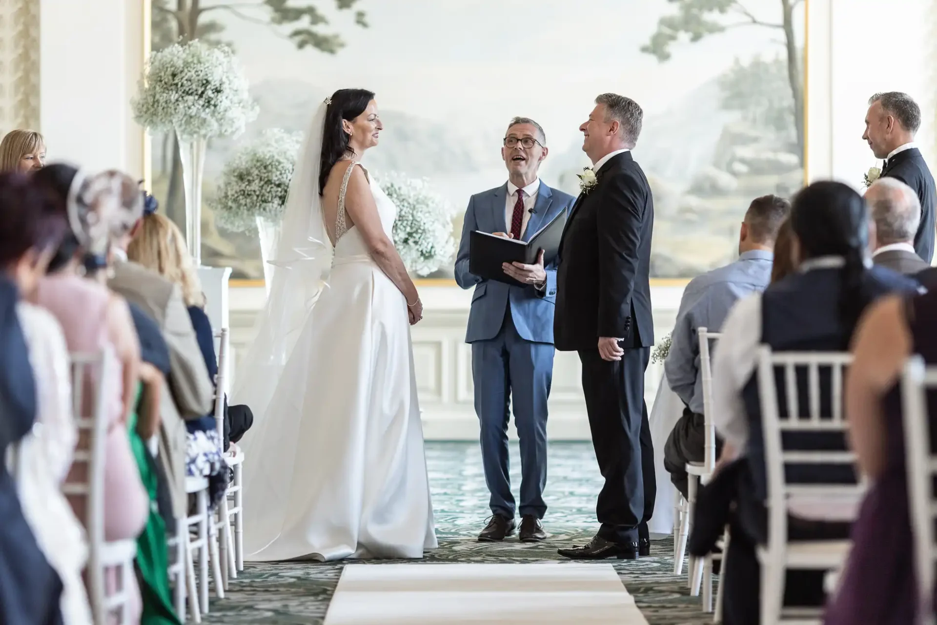 A bride and groom exchanging vows at a wedding ceremony indoors, with an officiant and guests watching.
