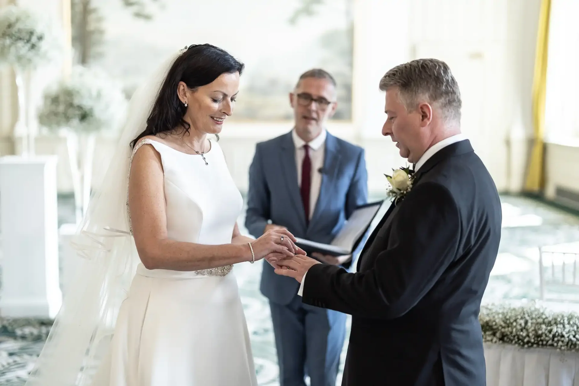 Bride in a white dress and groom in a black suit exchange rings during a wedding ceremony in a bright room, with an officiant standing in the background.