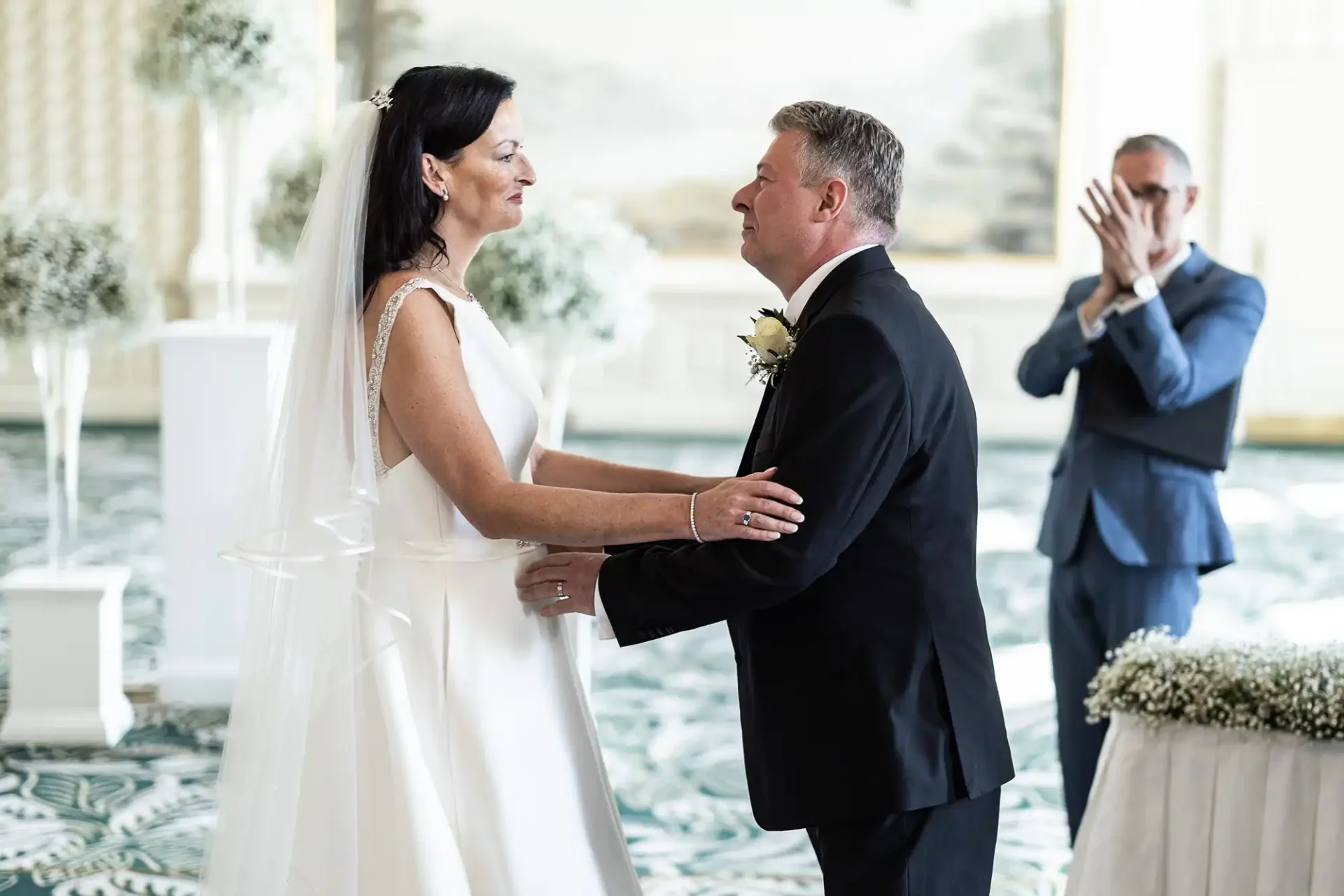 Bride and groom holding hands and smiling at each other during their wedding ceremony, with an emotional man applauding in the background.