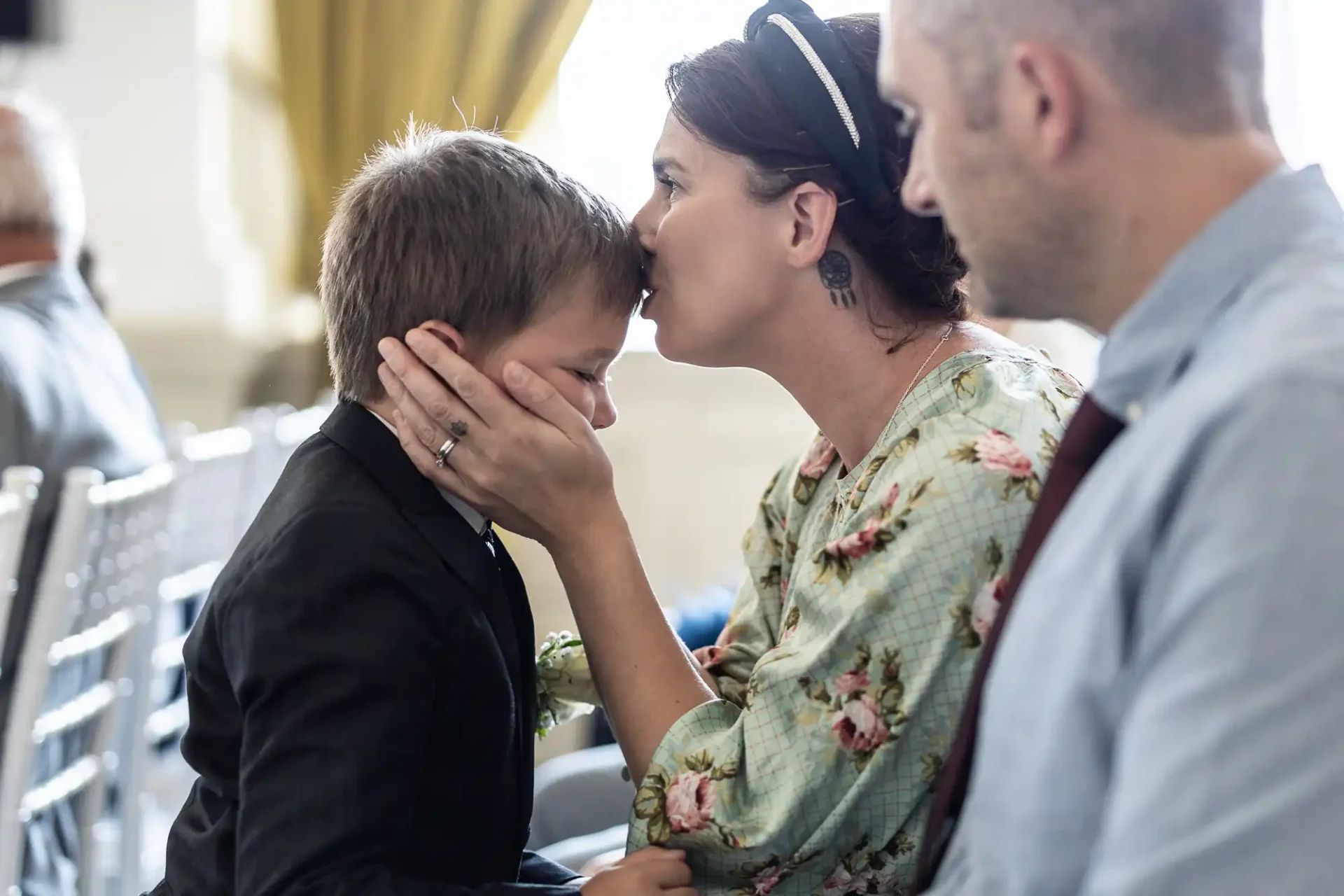 A woman in a floral dress kissing a young boy on the forehead at a formal event, with a man seated beside them.