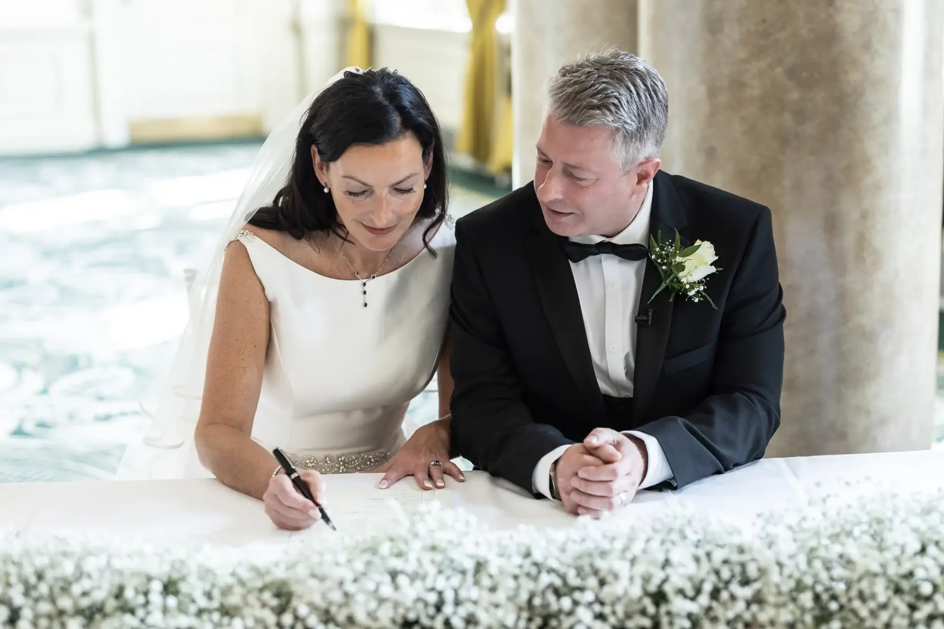 A bride in a white dress and a groom in a black tuxedo sign a document together at a table adorned with white flowers.