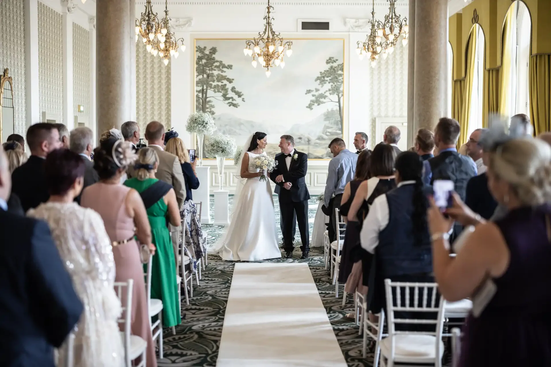A bride and groom exchanging vows at the front of an elegant room, surrounded by guests seated and standing, with ornate decor and large scenic murals on the walls.