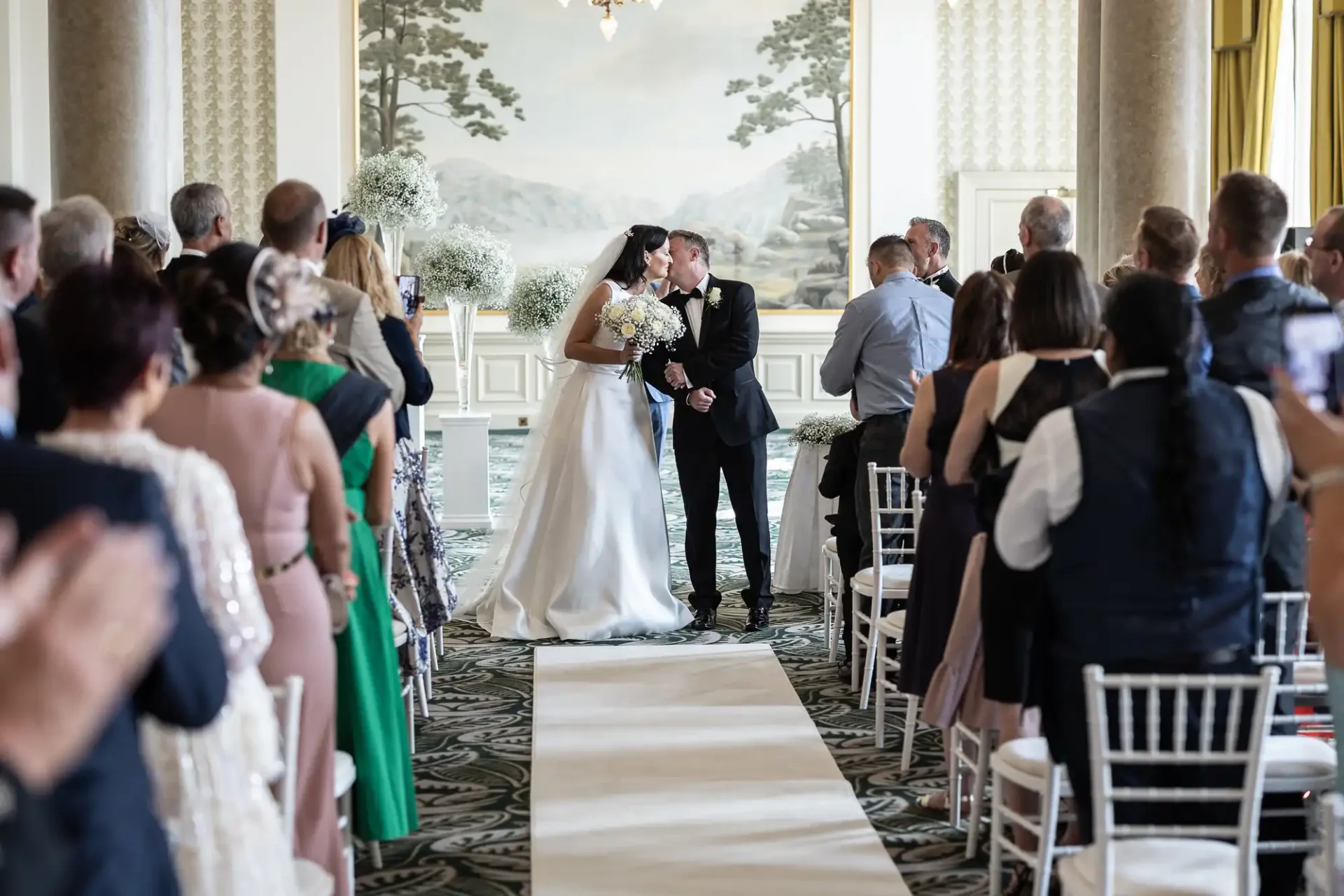 Bride and groom kissing at the altar in a grand room with large windows and guests standing around them, clapping.