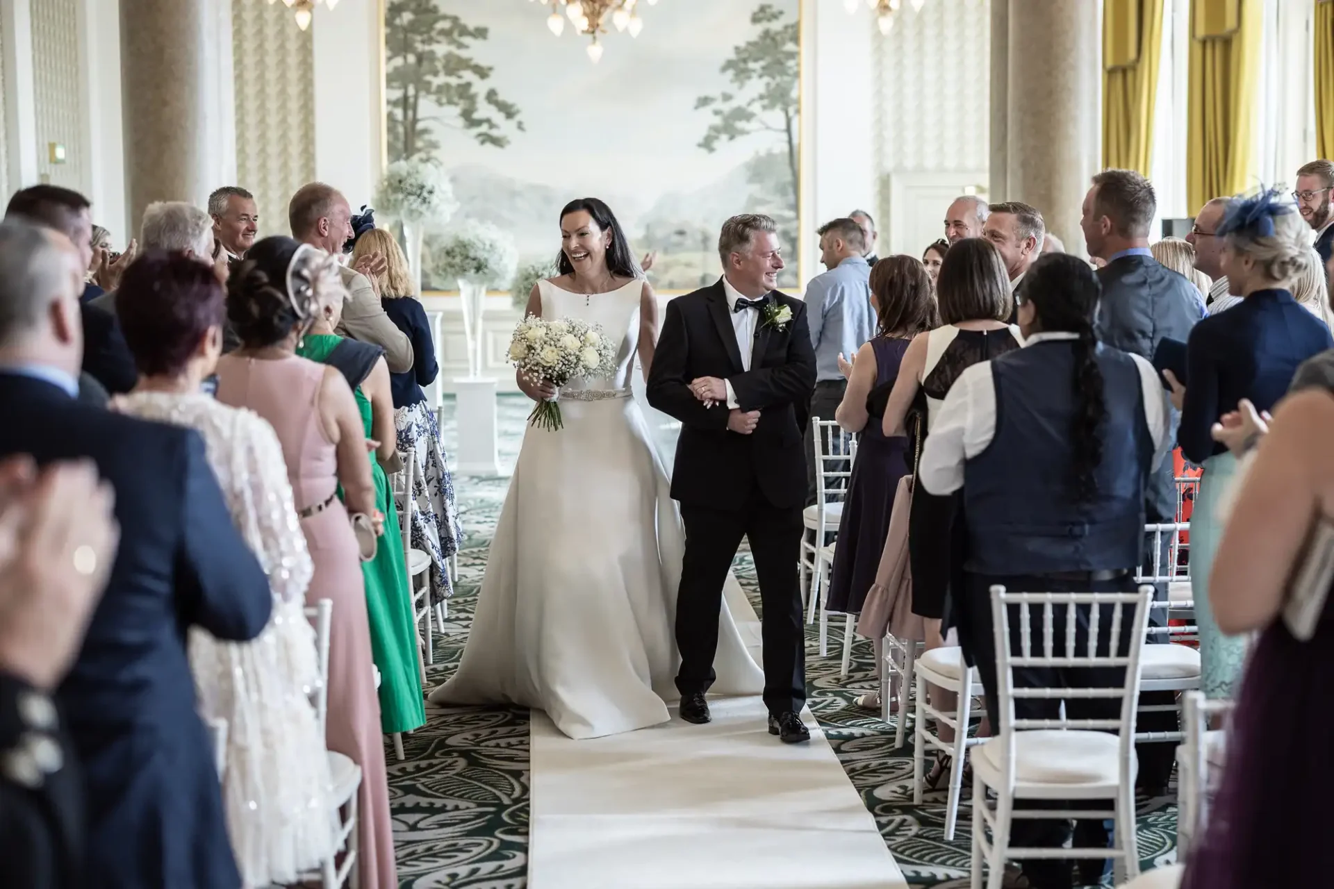 A bride and groom walk down the aisle, smiling, surrounded by applauding guests in an elegant room.