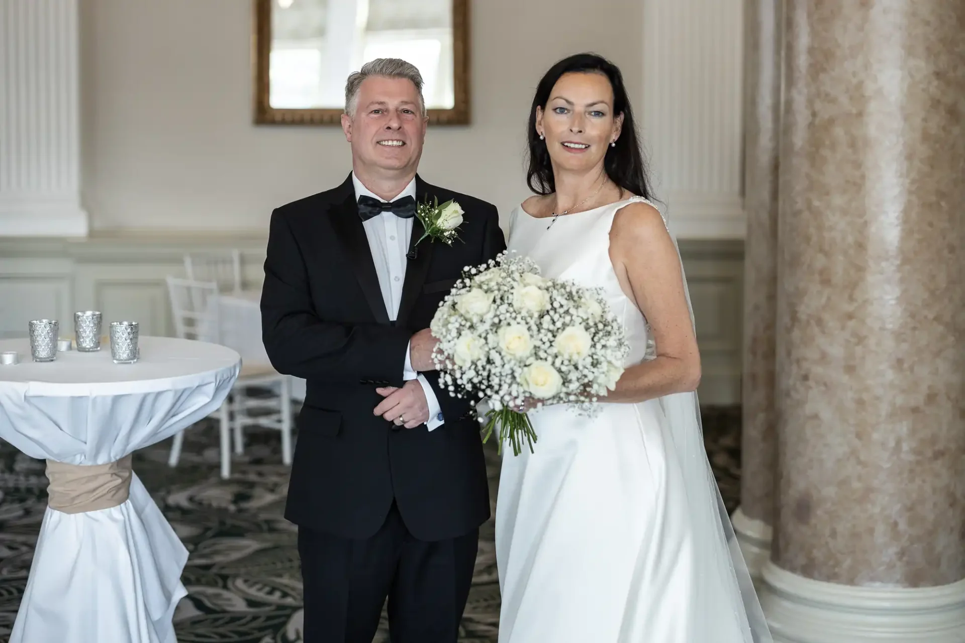 A bride and groom posing in a wedding venue, the bride holding a bouquet of white flowers, both smiling.
