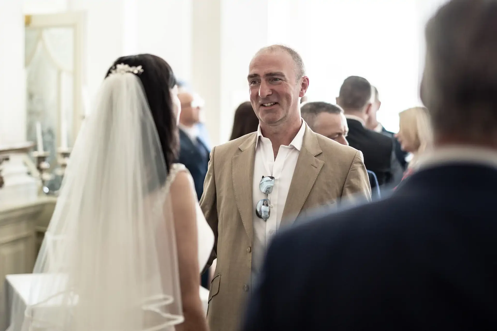 A smiling man in a beige suit at a wedding reception, conversing with guests including a bride in white.