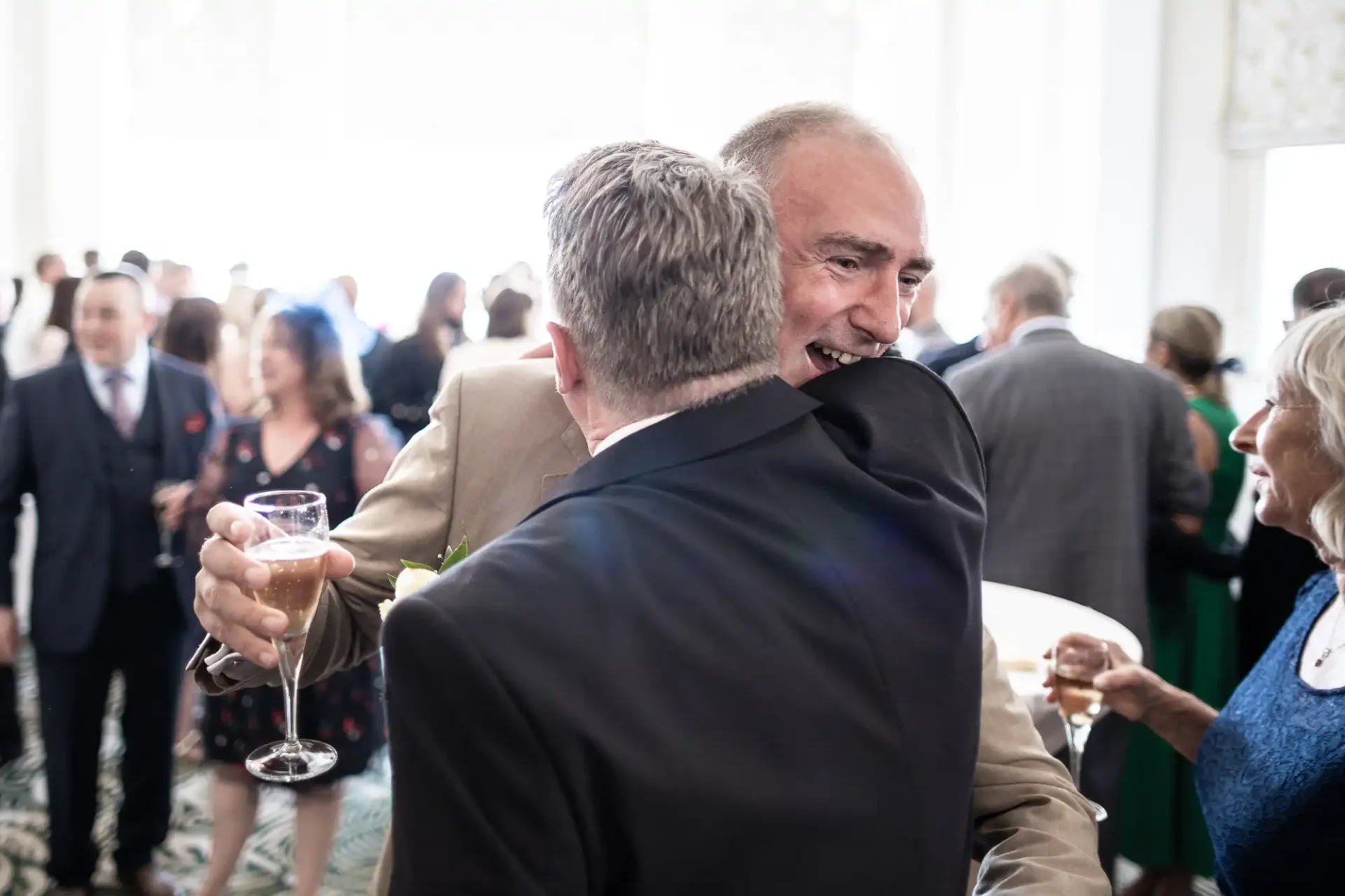 Two men embracing at a social event, one holding a glass of champagne, with other guests mingling in the background.