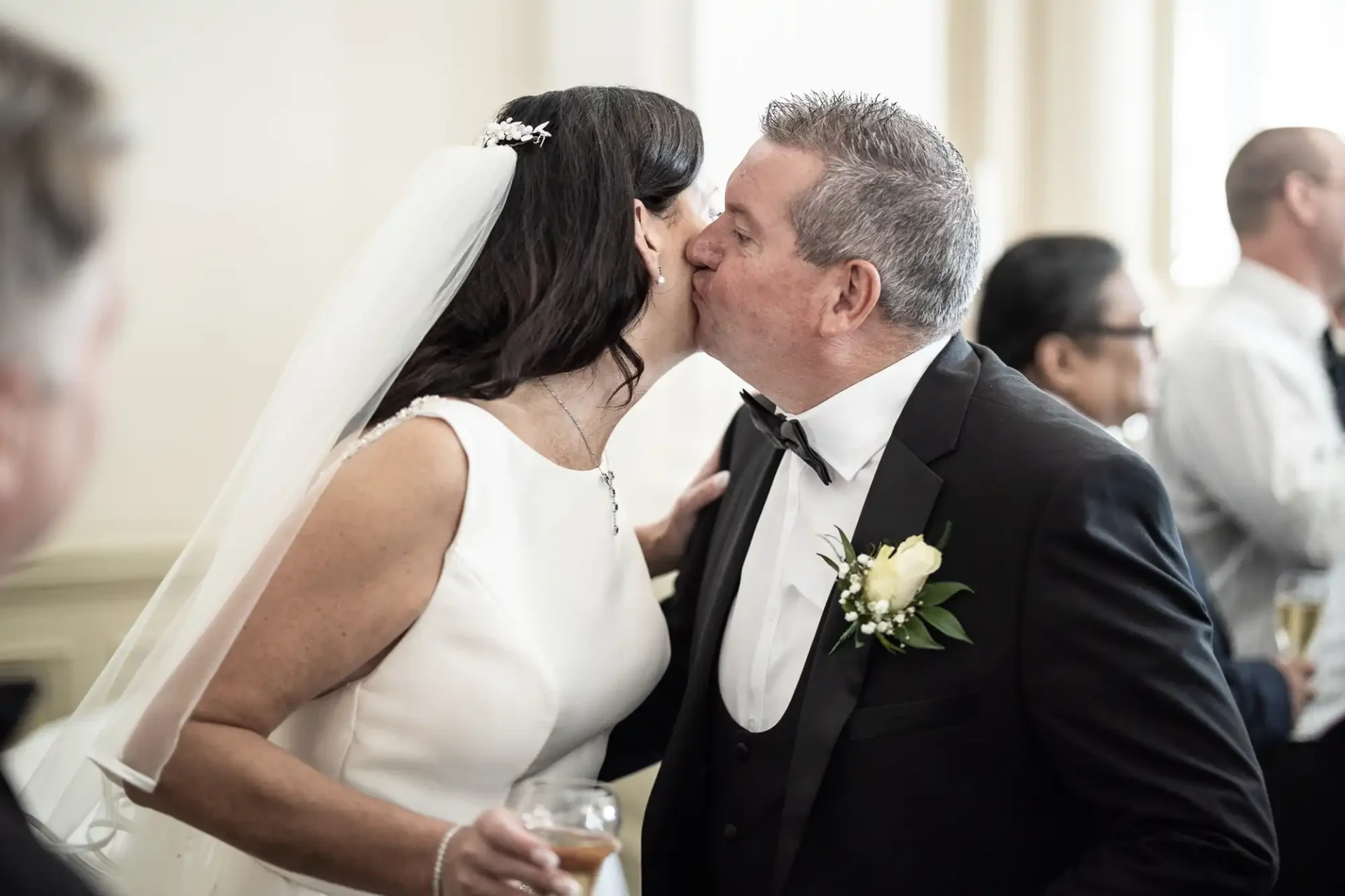 A bride and groom sharing a kiss at their wedding reception, with guests in the background.