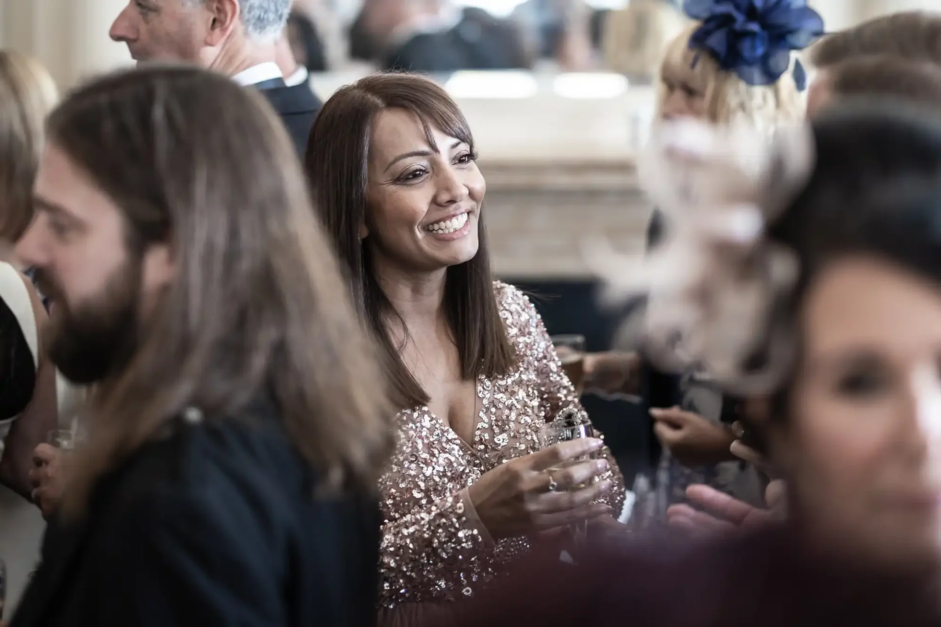 A woman in a sparkly dress smiling and conversing at a social gathering, surrounded by other guests in a softly blurred background.