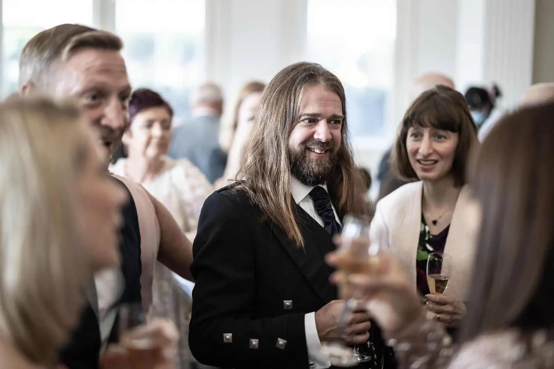A man with long hair smiling and holding a champagne glass at a social event, surrounded by guests engaged in conversations.