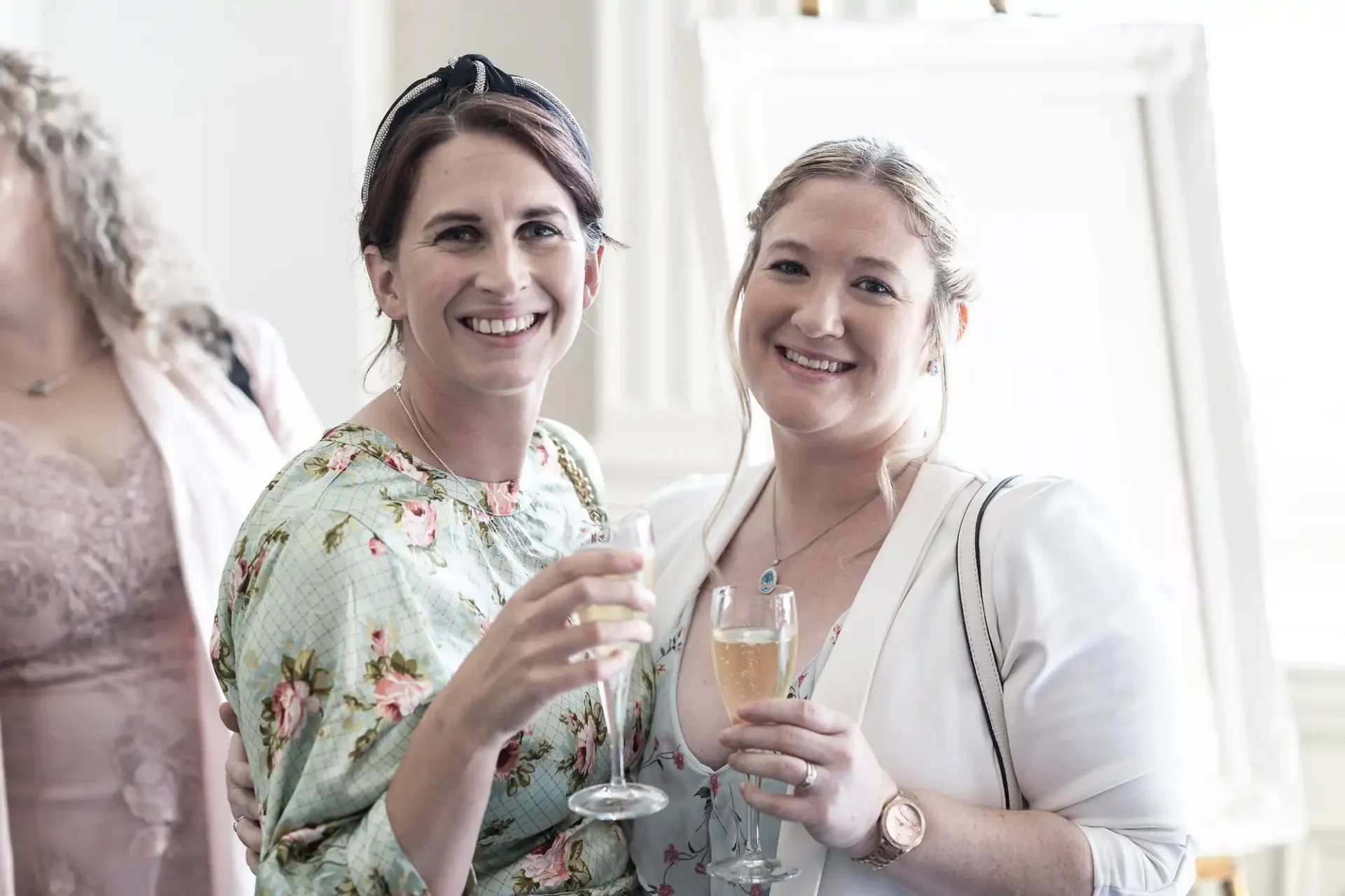 Two women smiling, holding champagne glasses at a social event, dressed in light casual elegant attire.