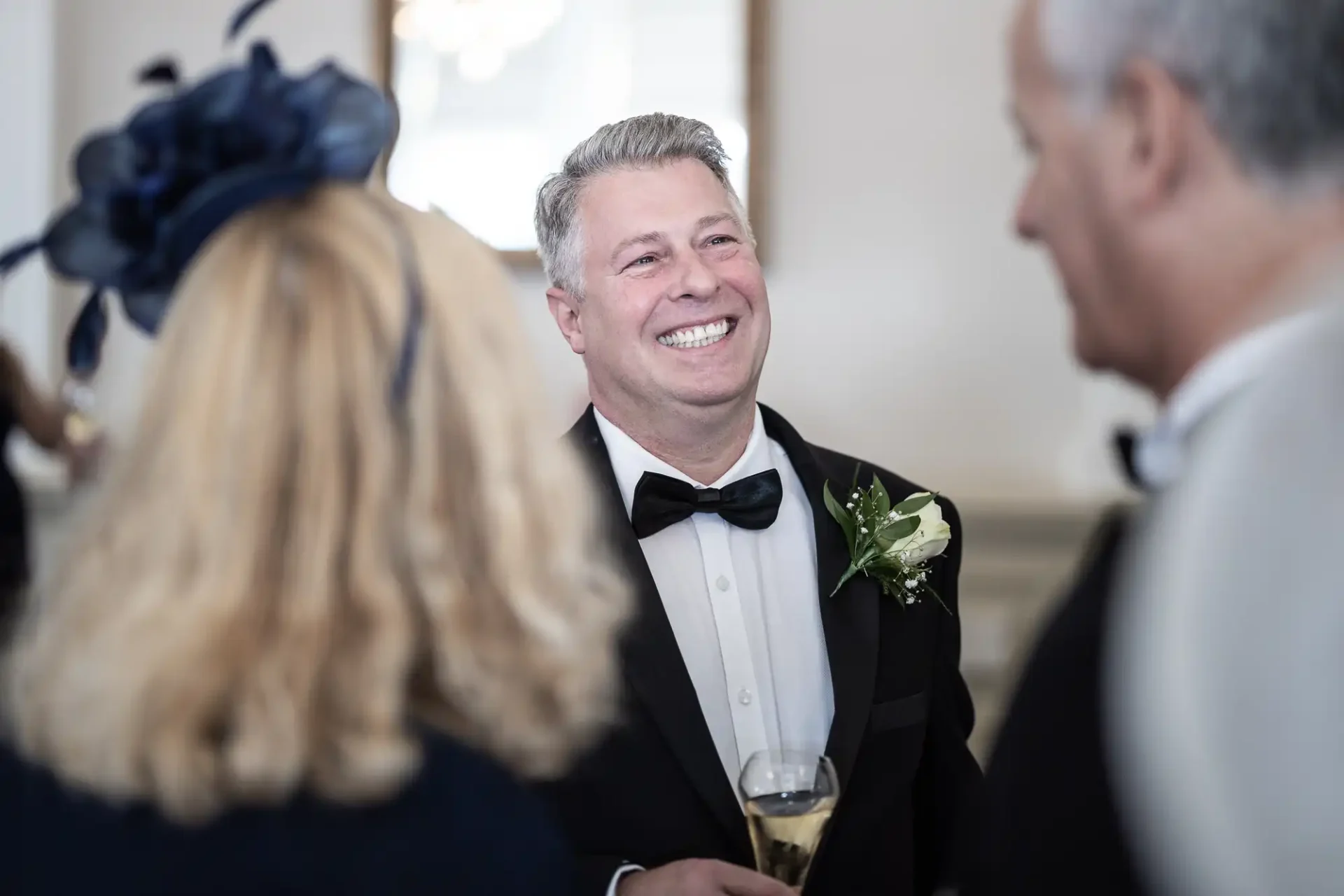 Man in a tuxedo with a boutonniere, holding a champagne flute, smiling while conversing with guests at a formal event.