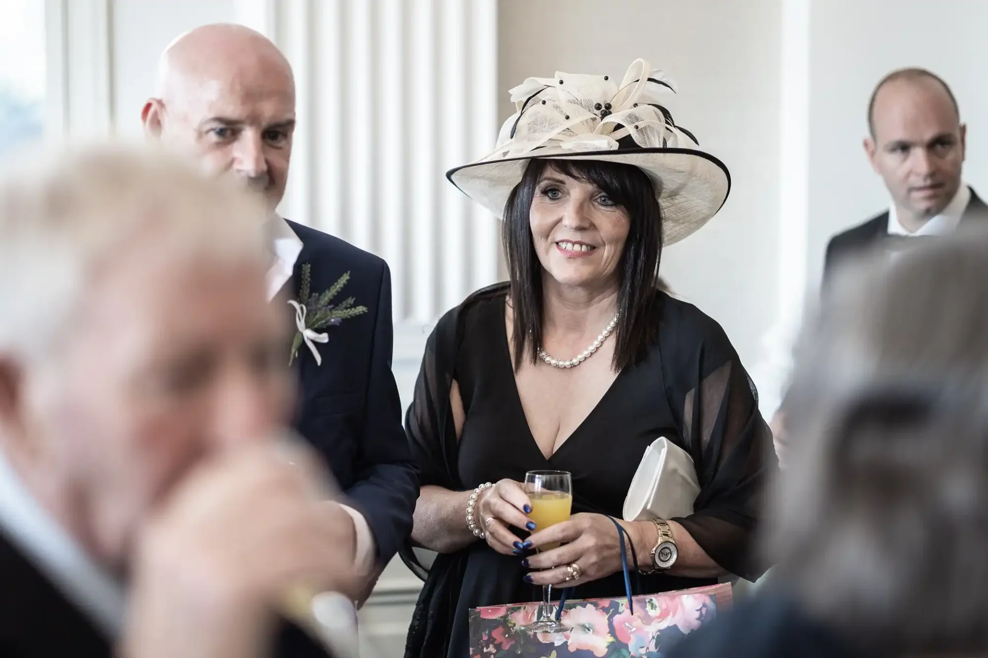 A woman in a black dress and a wide-brimmed hat holds a drink at a social gathering, with men in suits visible in the background.