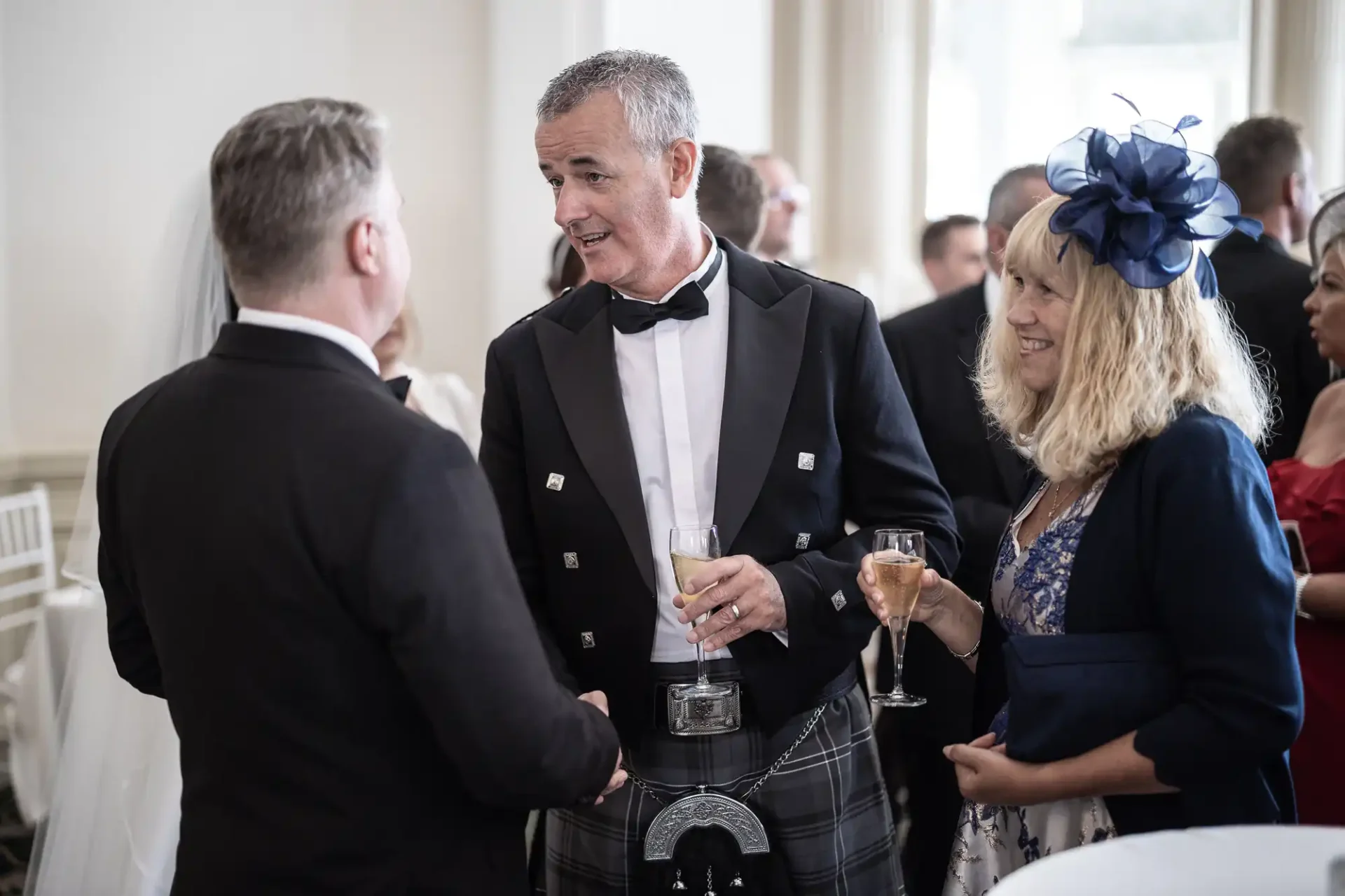 Two men and a woman engaging in conversation at a formal event, with one man dressed in a traditional kilt and holding a drink.