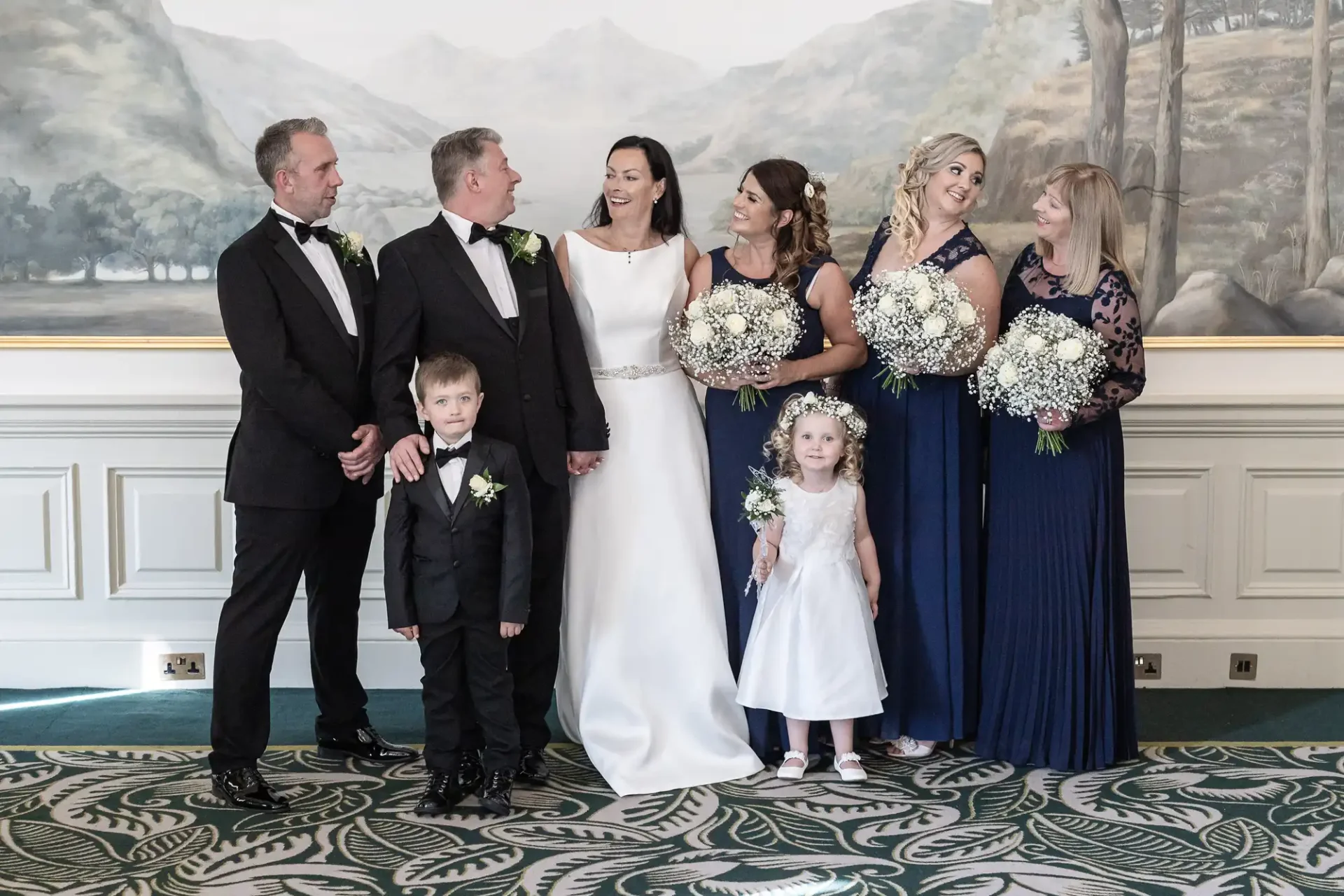 A wedding party posing indoors, featuring a bride and groom surrounded by four adults and two children, all dressed in formal attire.