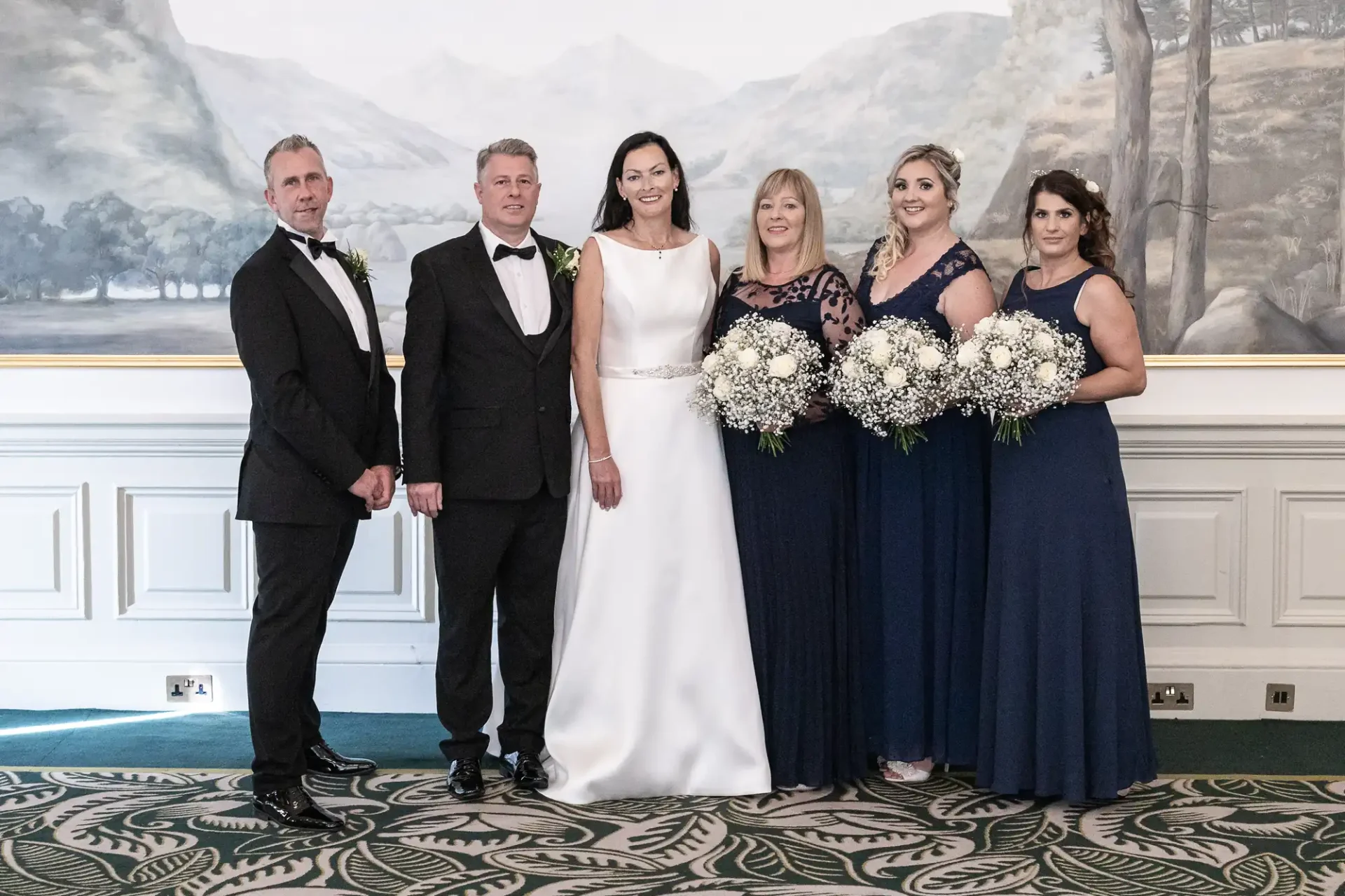 Wedding group photo featuring a bride and groom centered among three guests, two women in navy dresses and one man in a suit, all holding bouquets, in an elegant room.