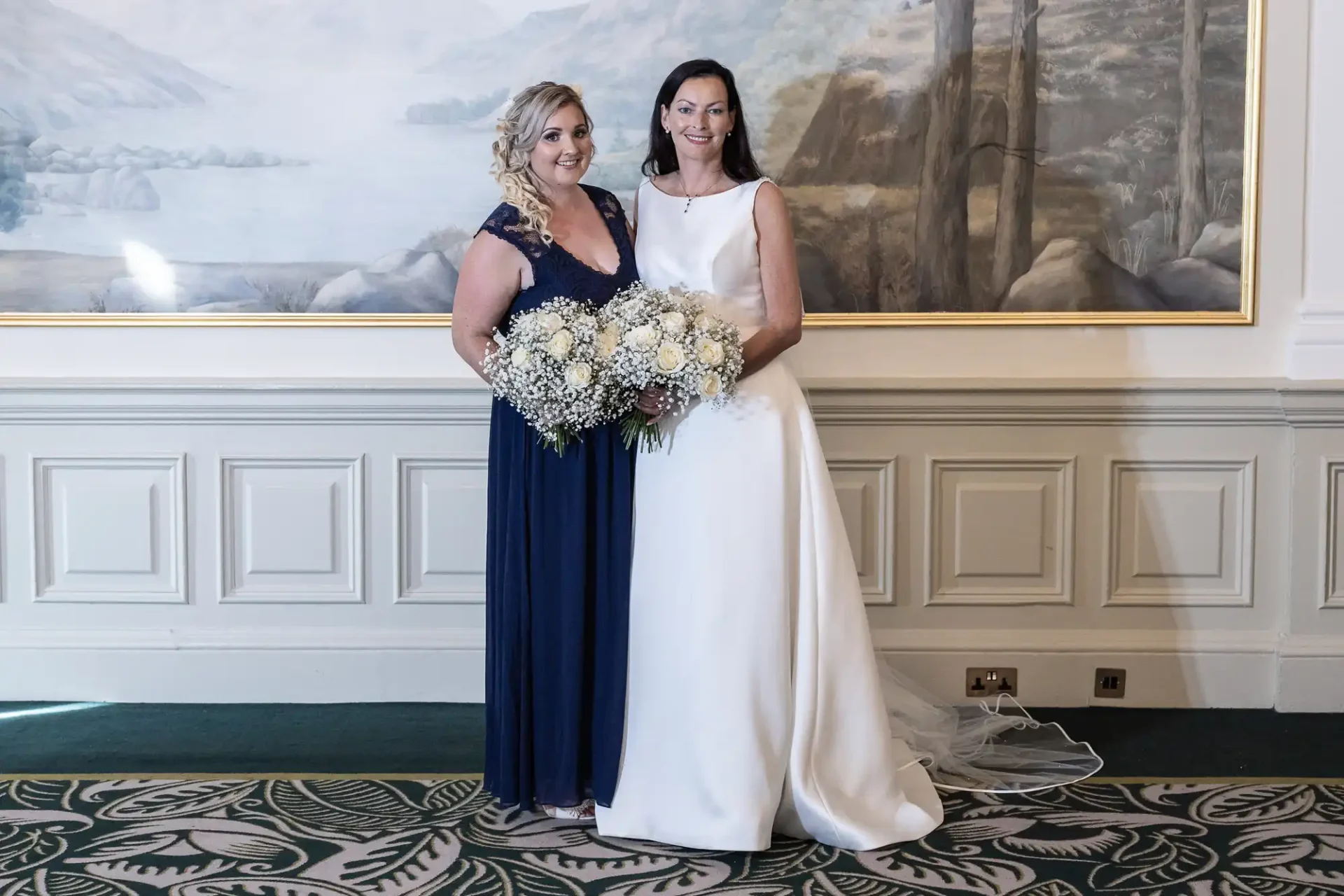 Two women in formal wear, one in a white wedding dress and the other in a navy gown, holding a bouquet, standing in front of a painting.