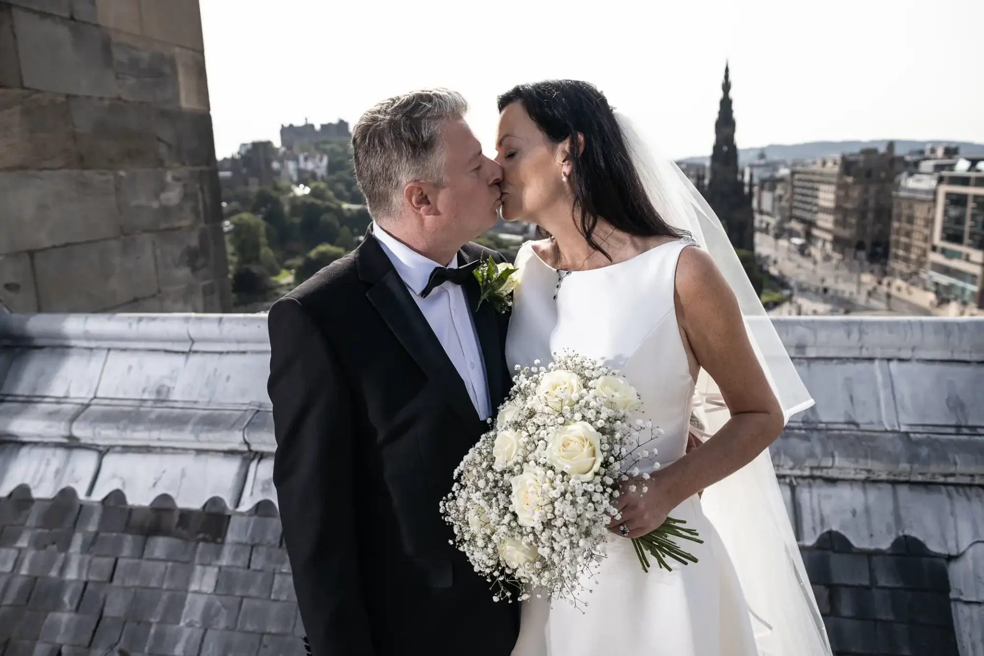 A bride and groom kissing on a rooftop with a cityscape in the background. the bride holds a bouquet of white flowers.