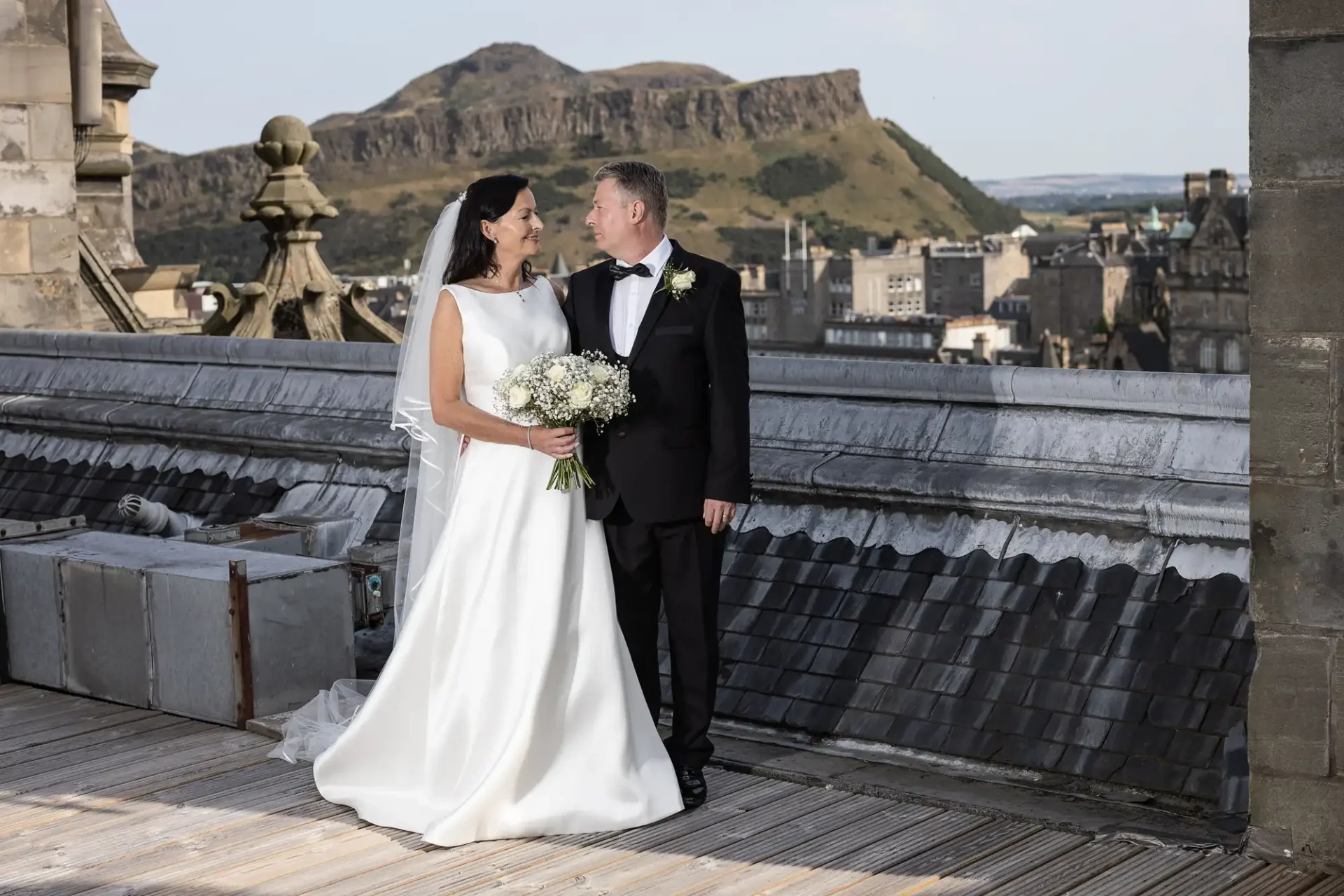 A bride and groom holding hands and smiling at each other on a rooftop, with a cityscape and mountains in the background.