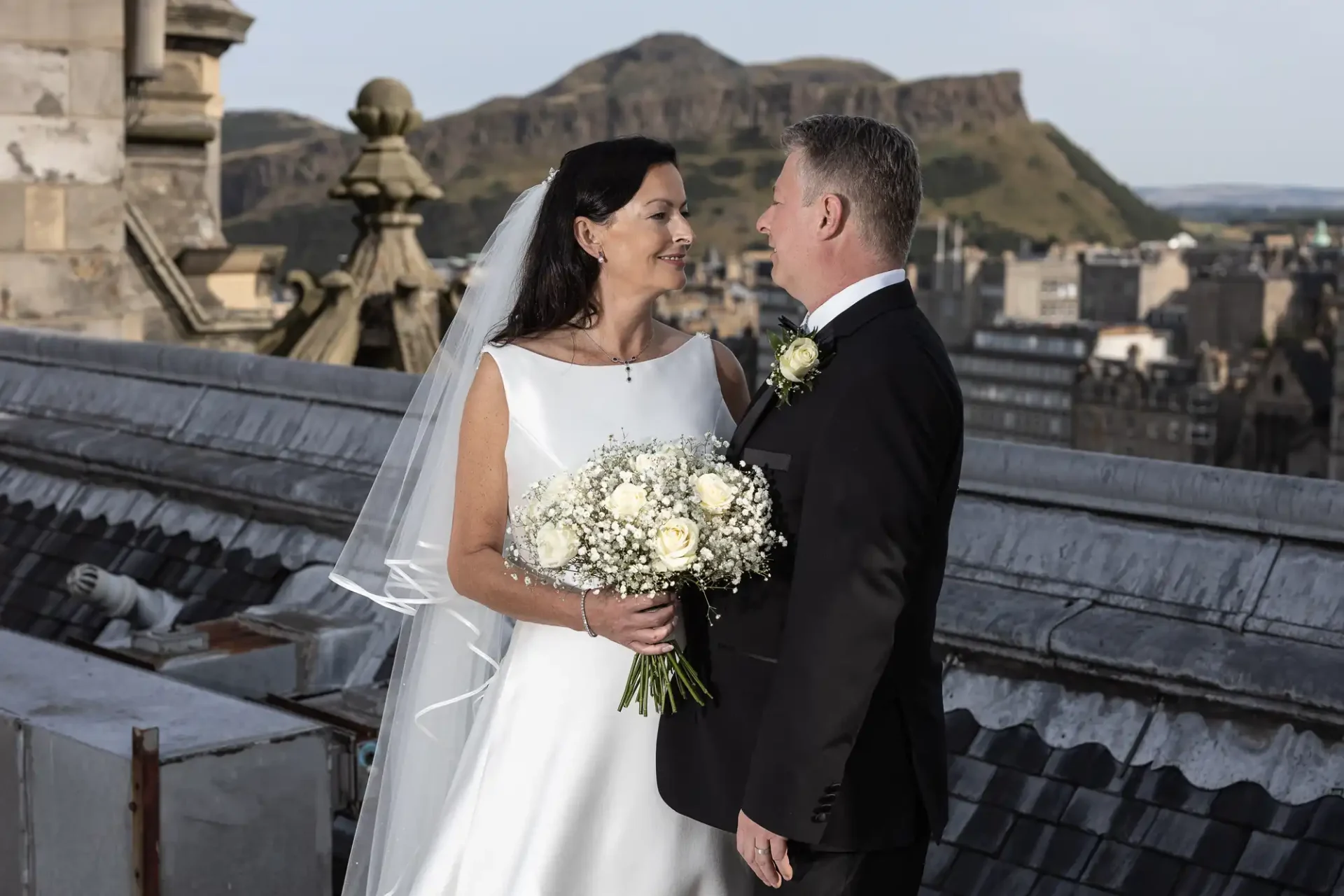 A bride and groom holding hands and smiling at each other on a rooftop with a cityscape and hills in the background.