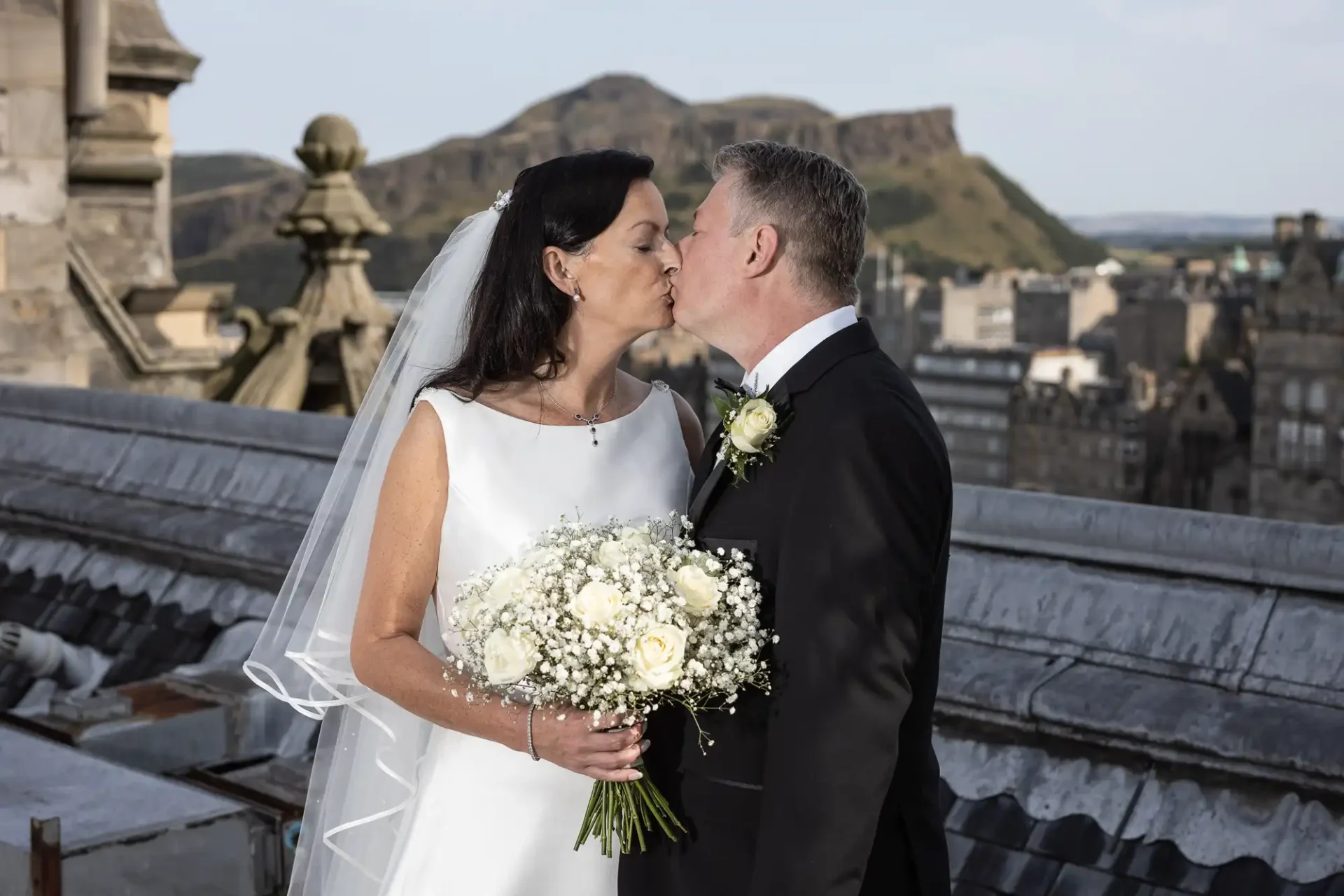 A bride and groom kissing on a rooftop, with a large bouquet and a cityscape featuring a hill in the background.