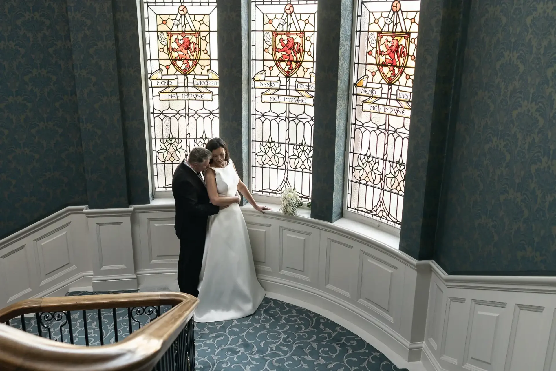 A bride and groom embrace on a staircase with ornate stained-glass windows in the background.