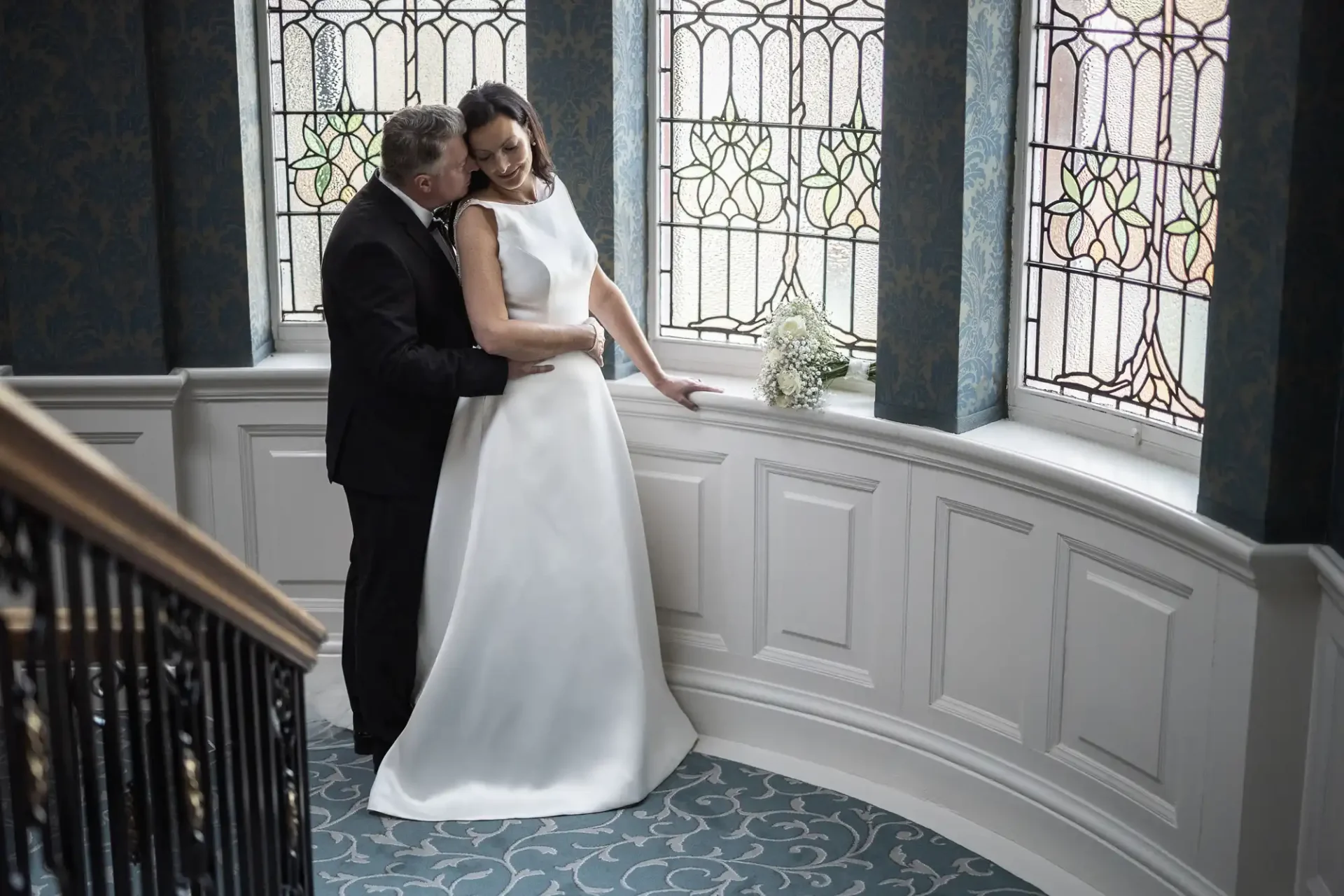 A newlywed couple embracing near a window with stained glass panels, the bride in a white gown and the groom in a suit.