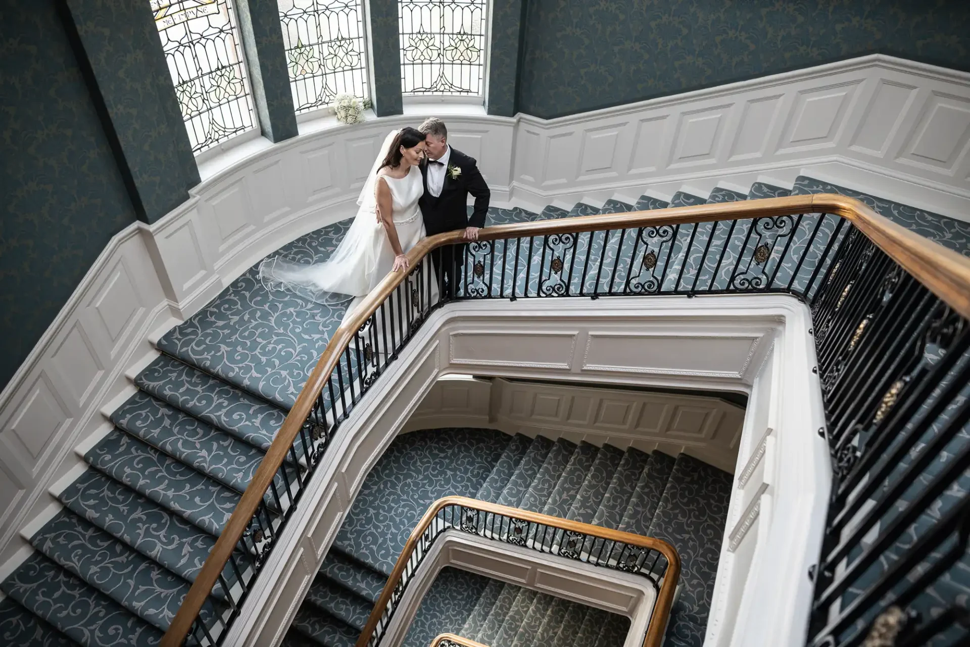A bride and groom sharing an intimate moment on a grand staircase with ornate railings and elegant wallpaper, viewed from above.