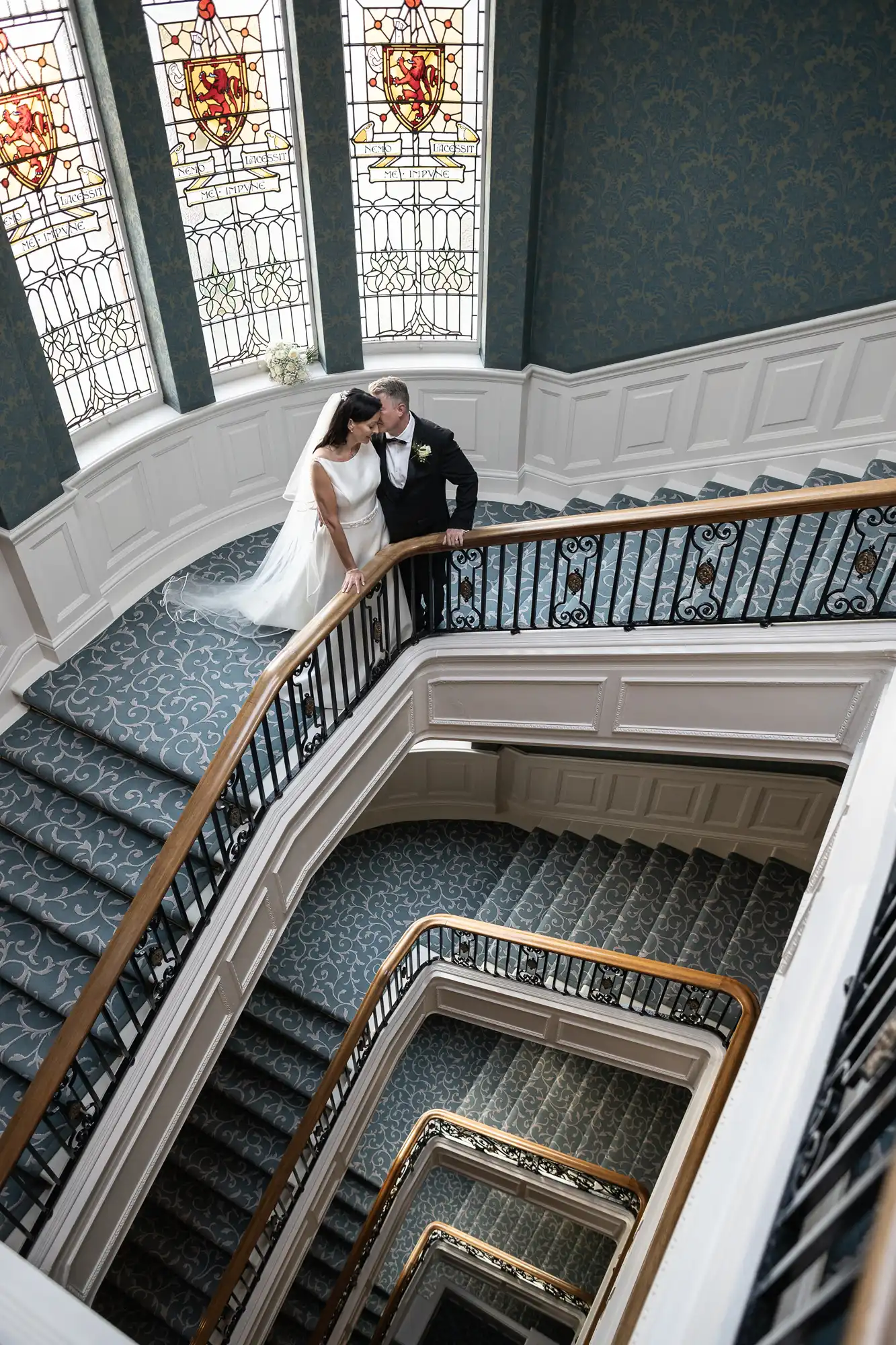 A bride and groom share an intimate moment on a spiraling staircase with ornate railings and stained glass windows in a grand building.