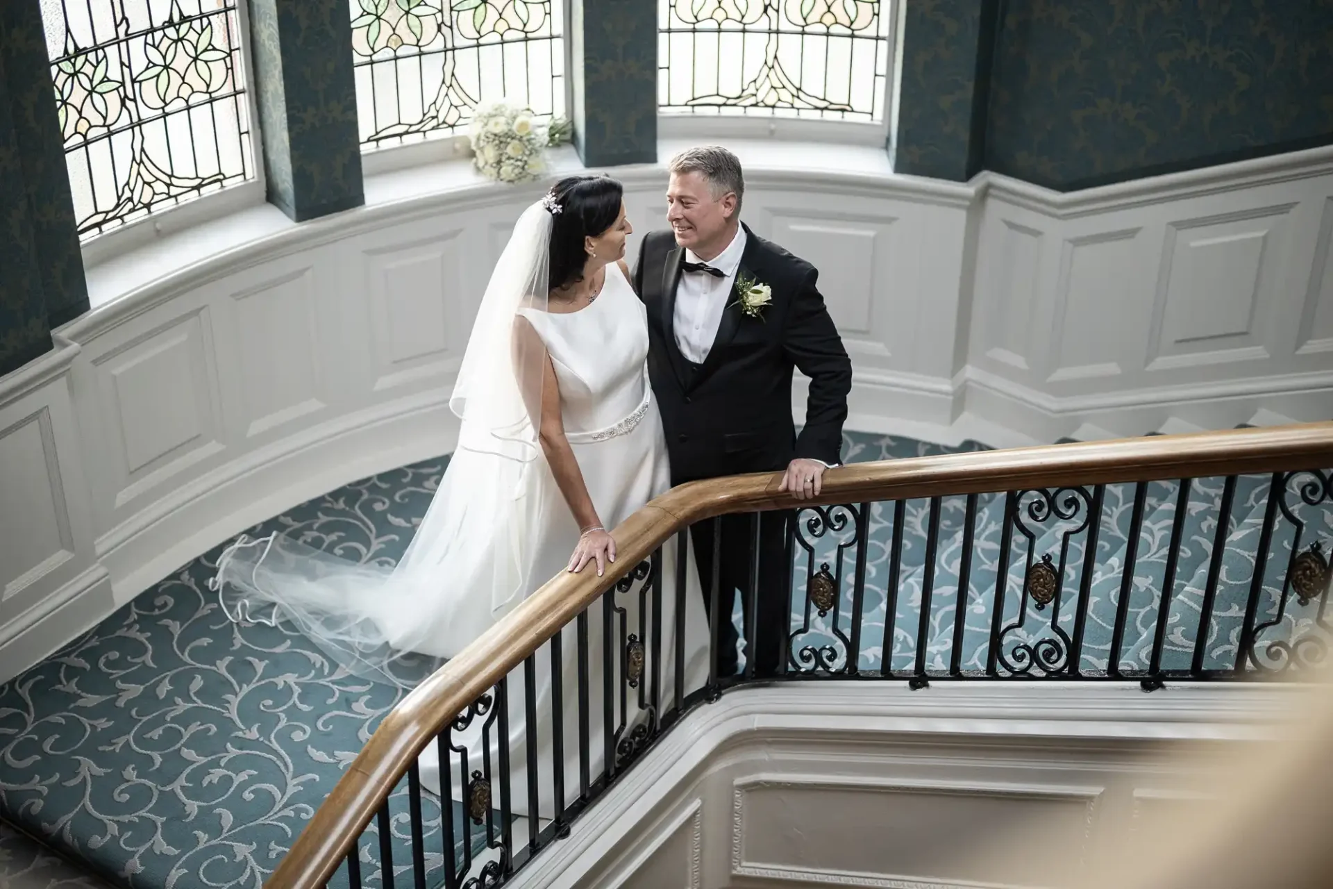 A bride and groom stand on an ornate staircase with stained glass windows, looking at each other affectionately.