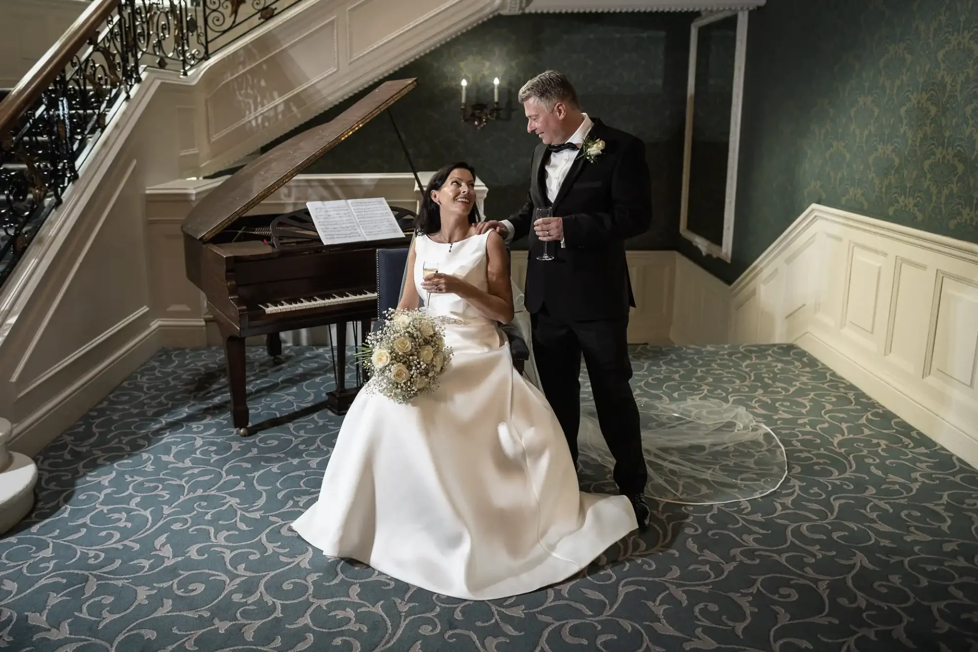 Bride and groom smiling at each other beside a piano in an elegant room, bride seated with a bouquet, groom standing with a glass in hand.