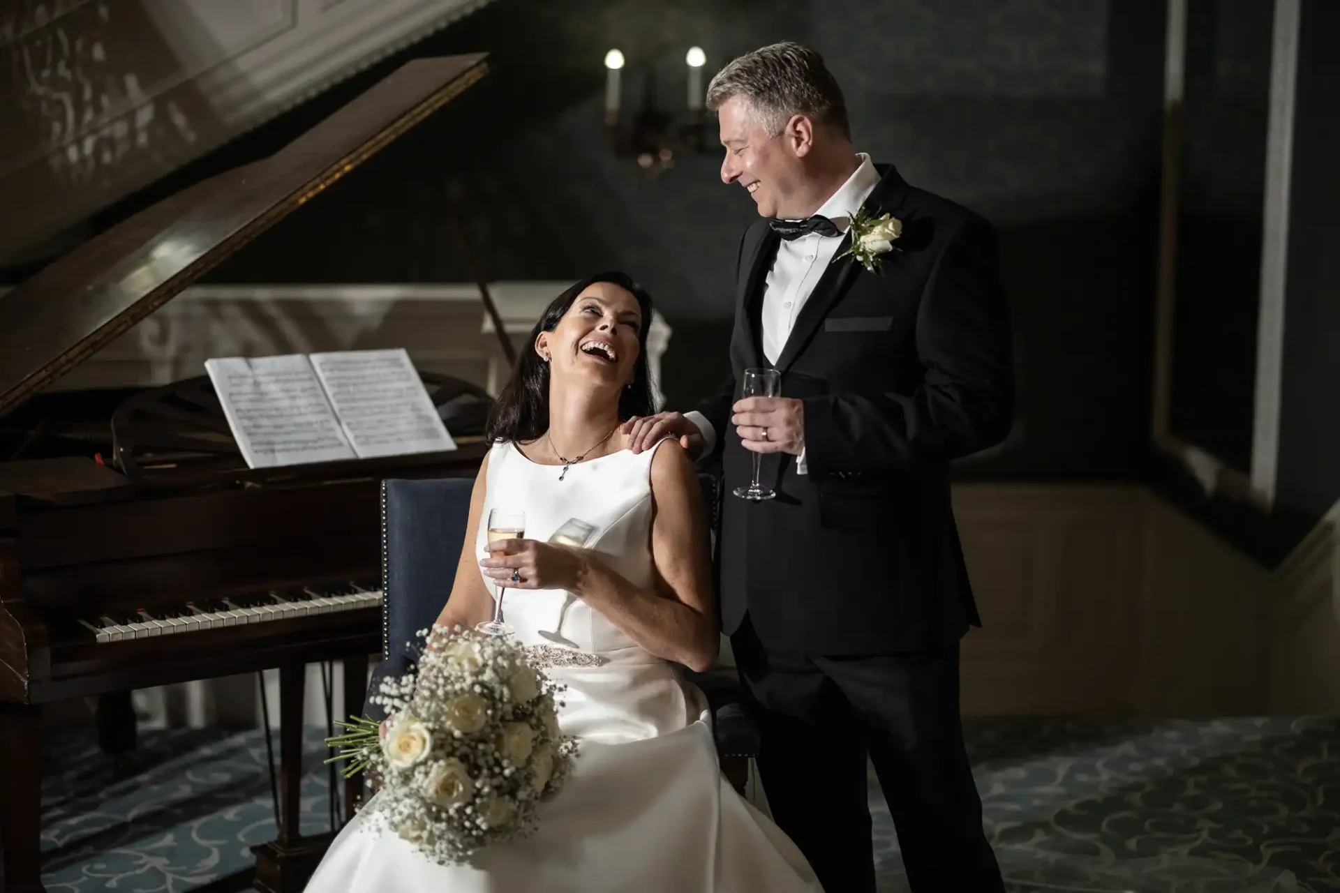 A bride and groom laughing together, holding drinks, beside a grand piano in an elegant room.