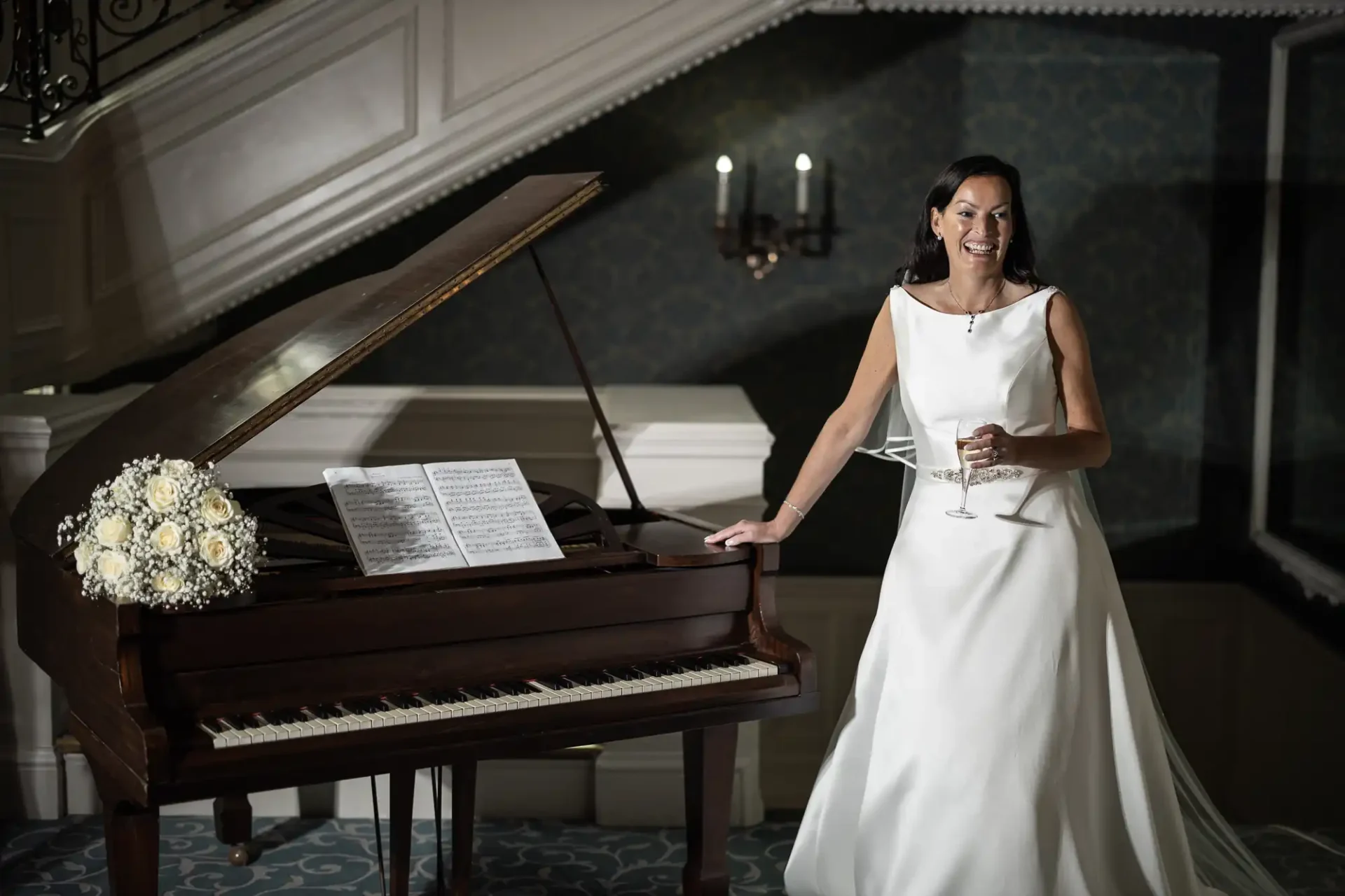 A smiling woman in a white wedding dress stands beside an open grand piano, holding a wine glass, in an elegant room.