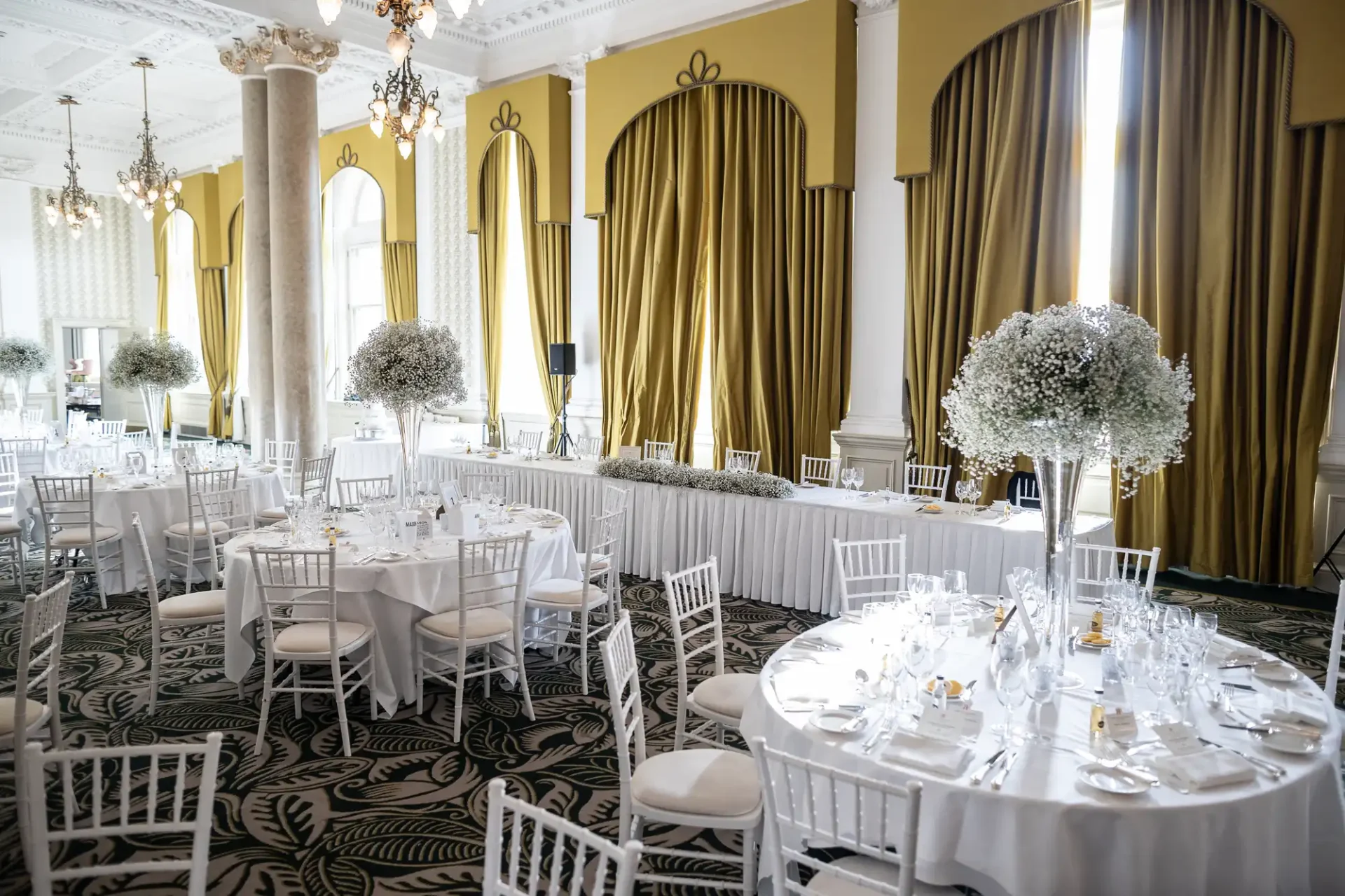 Elegant dining room setup for a wedding, featuring white chairs, round tables with floral centerpieces, and draped golden curtains.